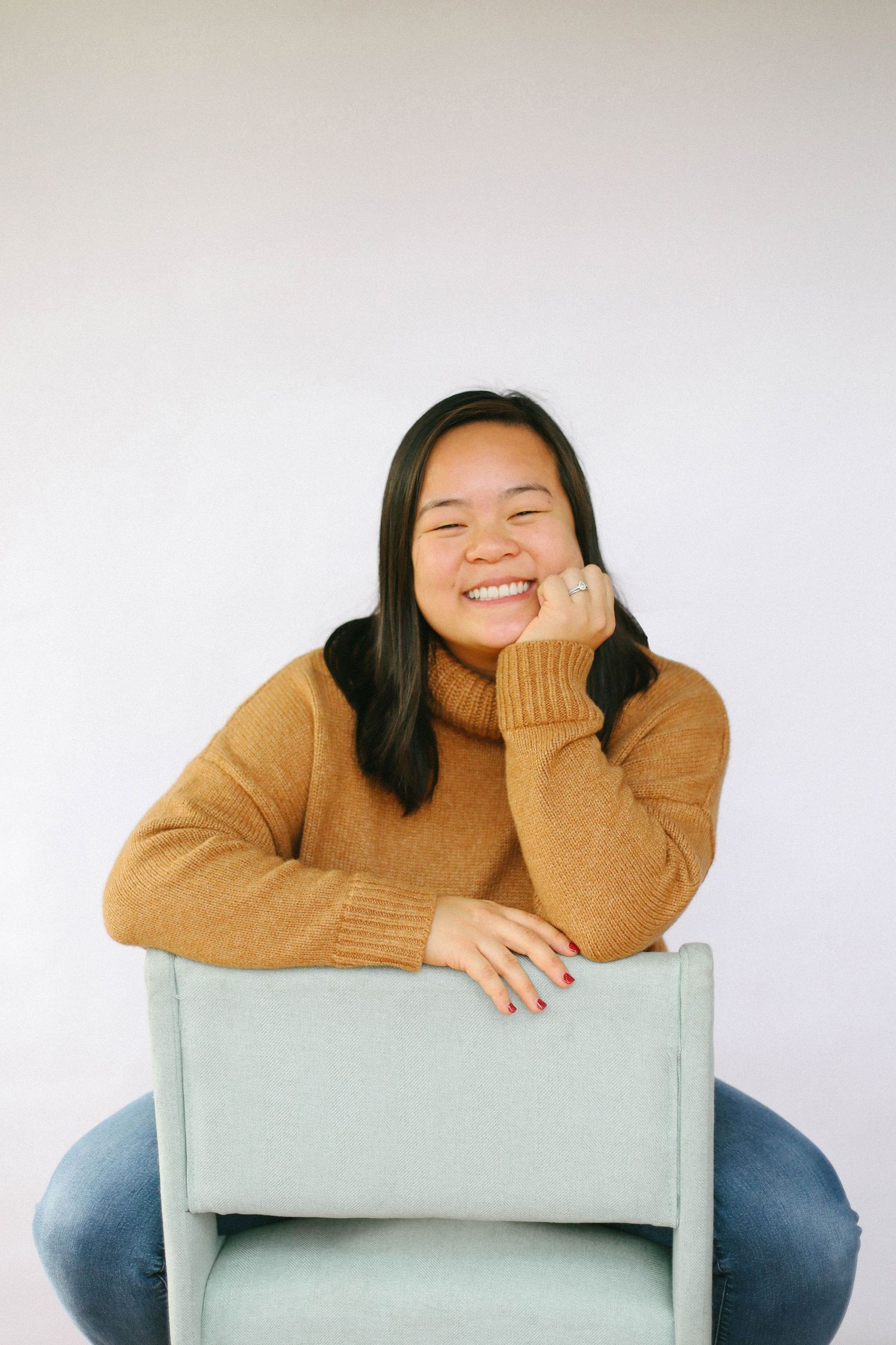 Smiling young Asian woman in a camel turtleneck sweater sitting relaxed in a mint chair against a white background.