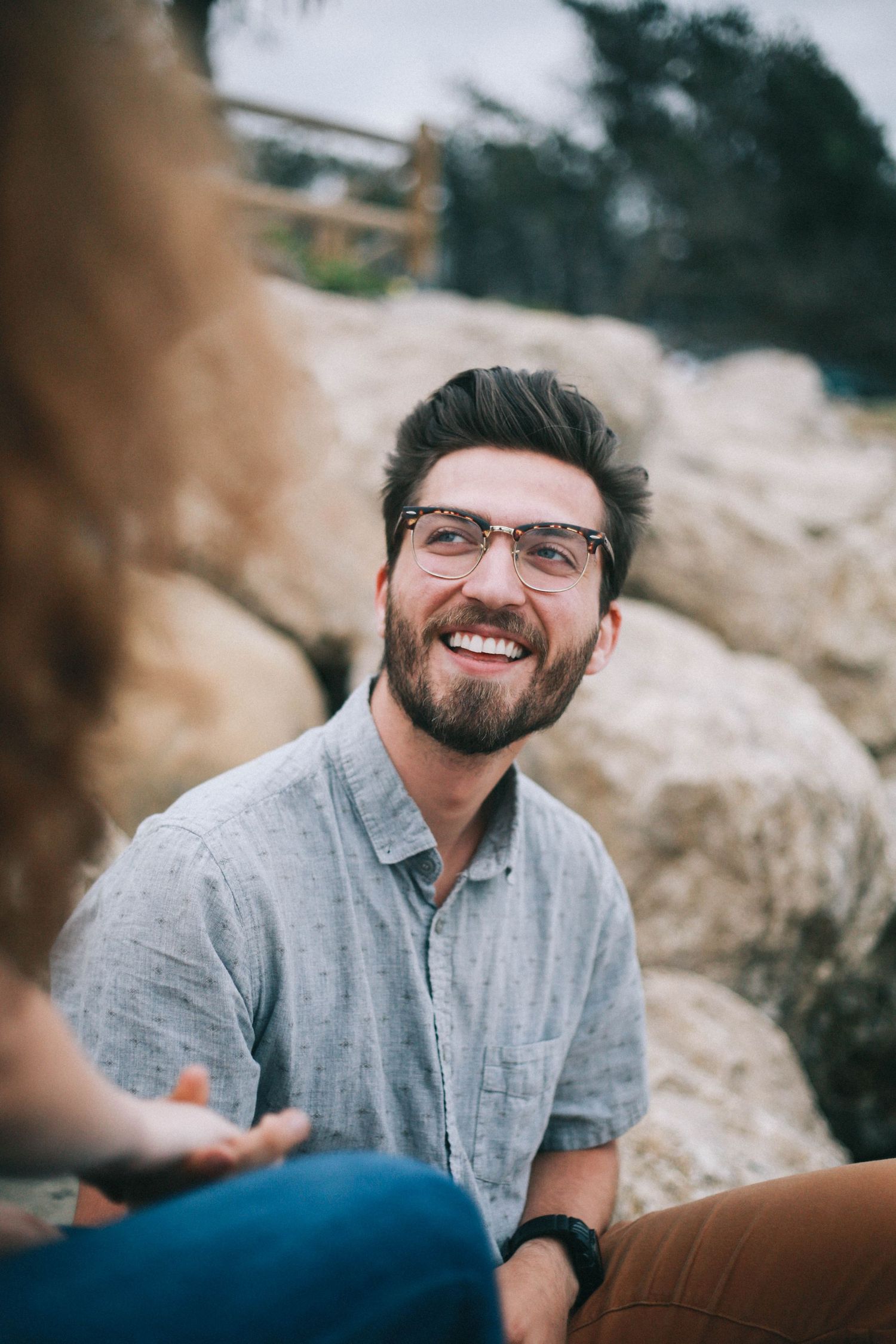 Smiling bearded man with glasses sits on rocks holding hands with his partner outdoors.