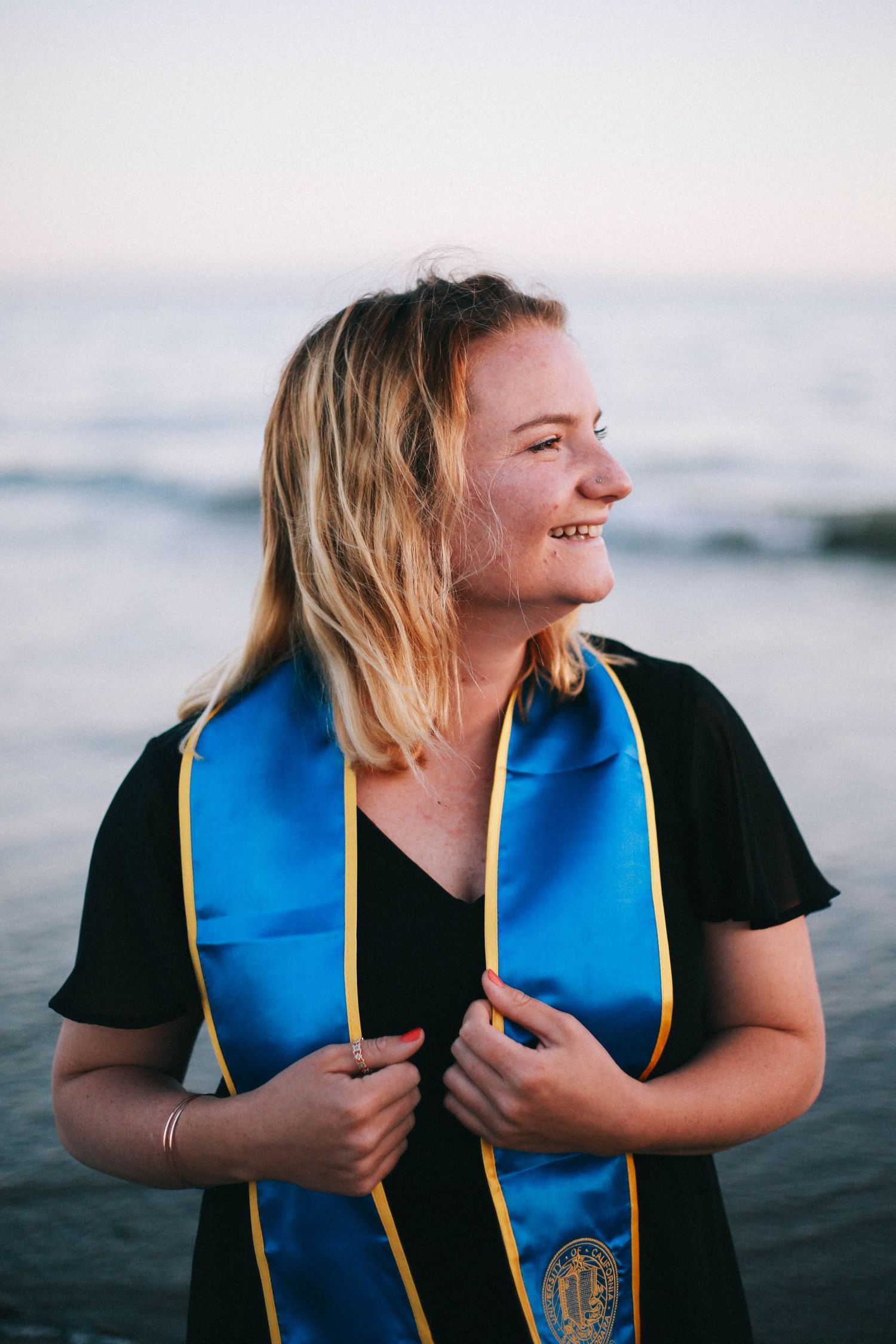 Smiling blonde woman wearing a blue and gold UCSB graduation stole at the beach during sunset.
