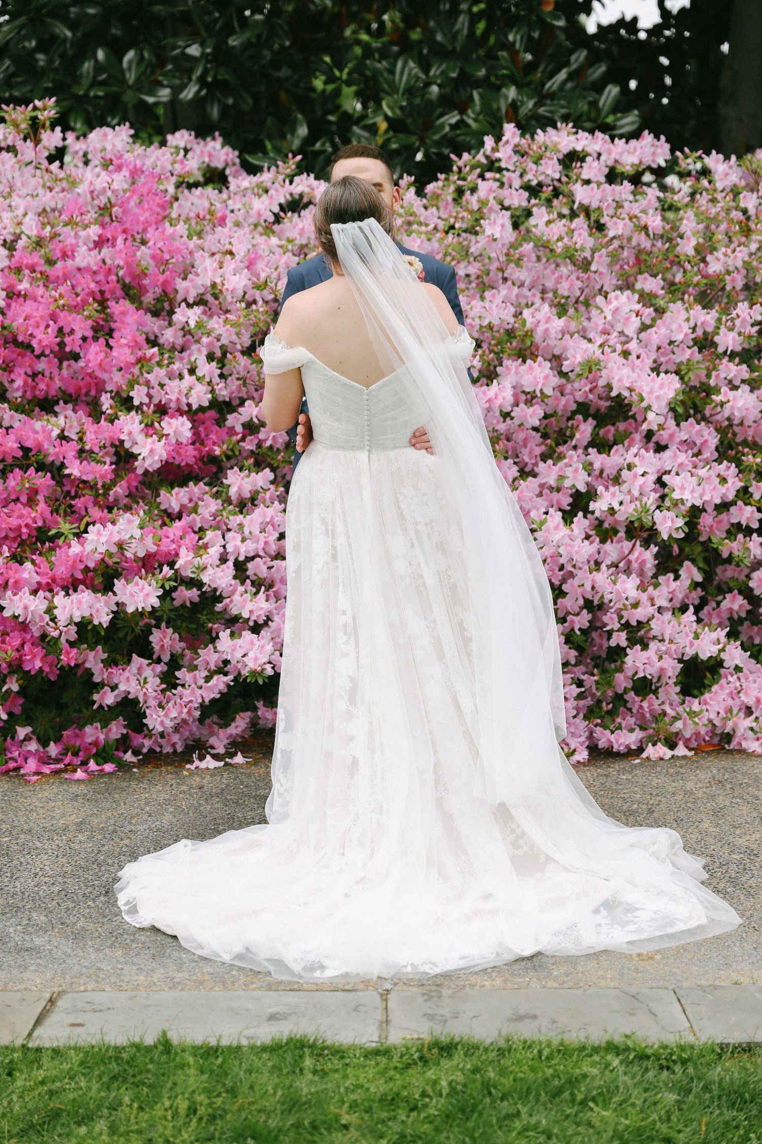 Bride in off-shoulder lace gown with veil stands before vibrant pink azalea blooms with groom.