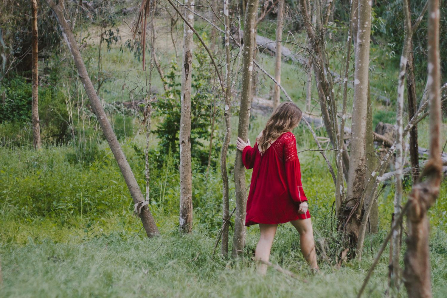 Woman in red dress walking through sparse woodland with green undergrowth.