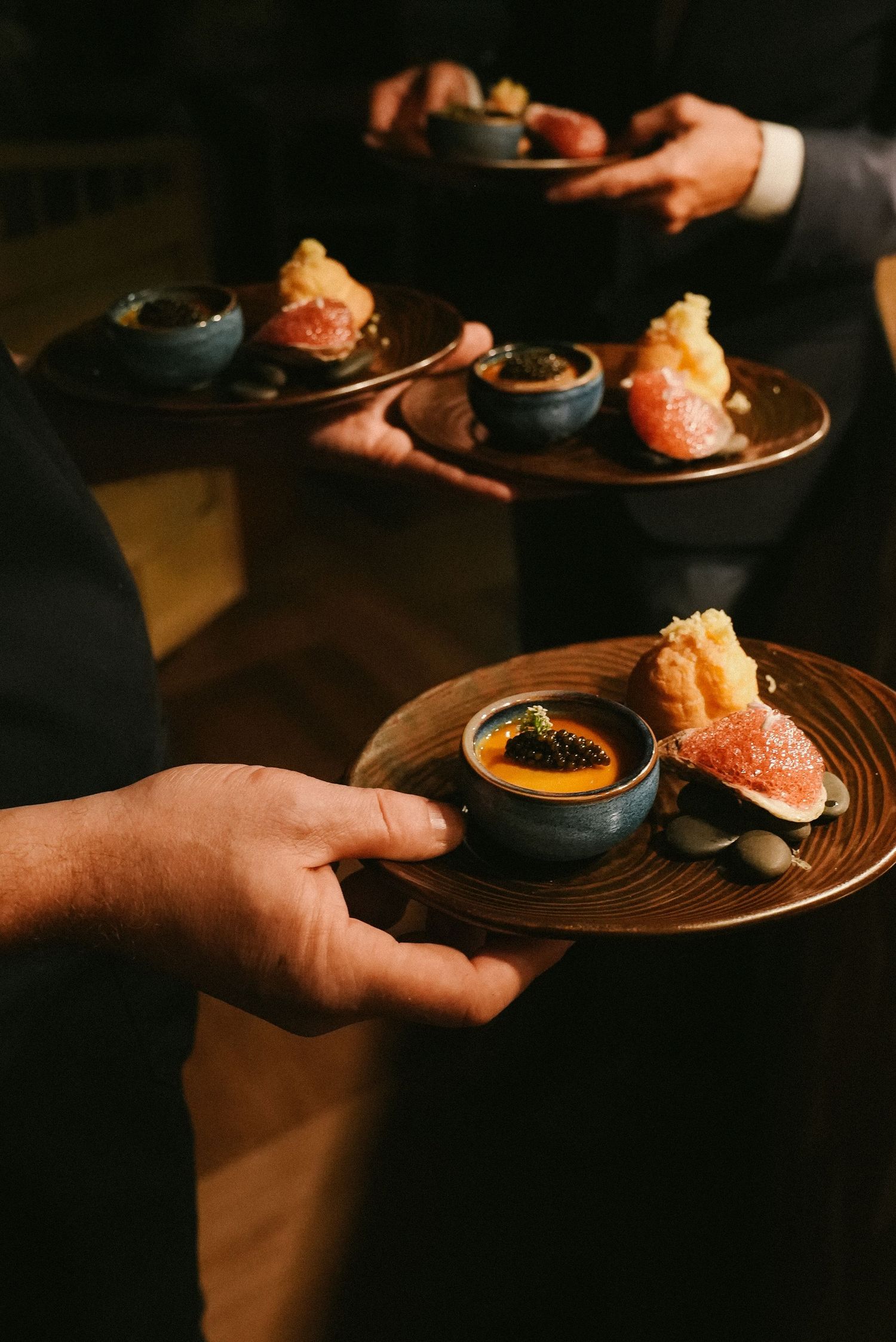 Waiters carrying elegant bronze trays with gourmet amuse-bouche featuring caviar and tempura bites.