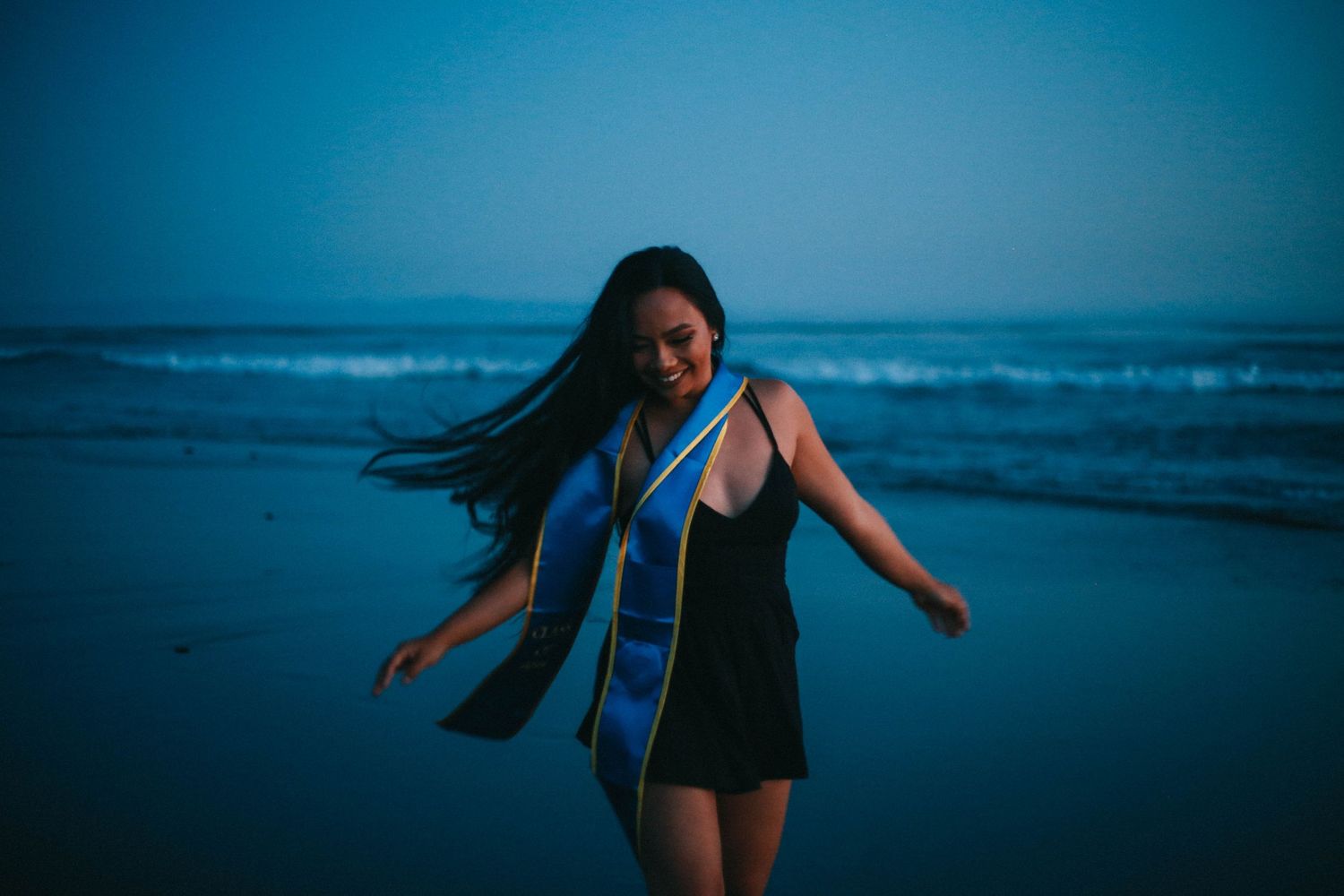 Young woman in black dress with blue graduation stole dancing on beach at twilight.