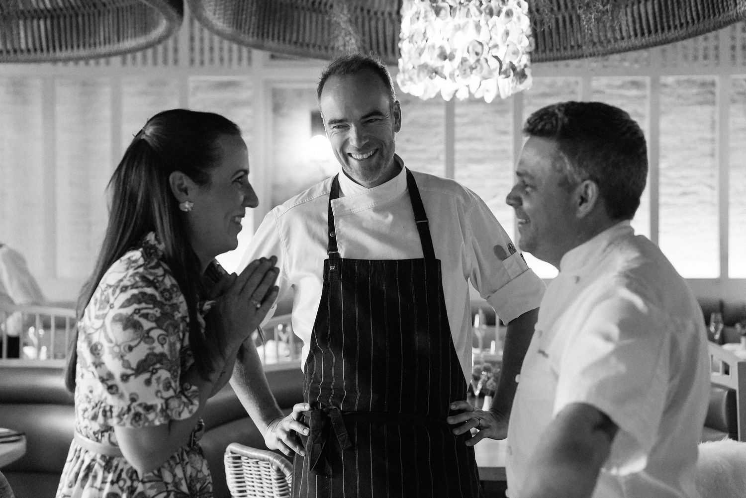 Two chefs in white uniforms chat warmly with a female guest in a stylish restaurant interior.