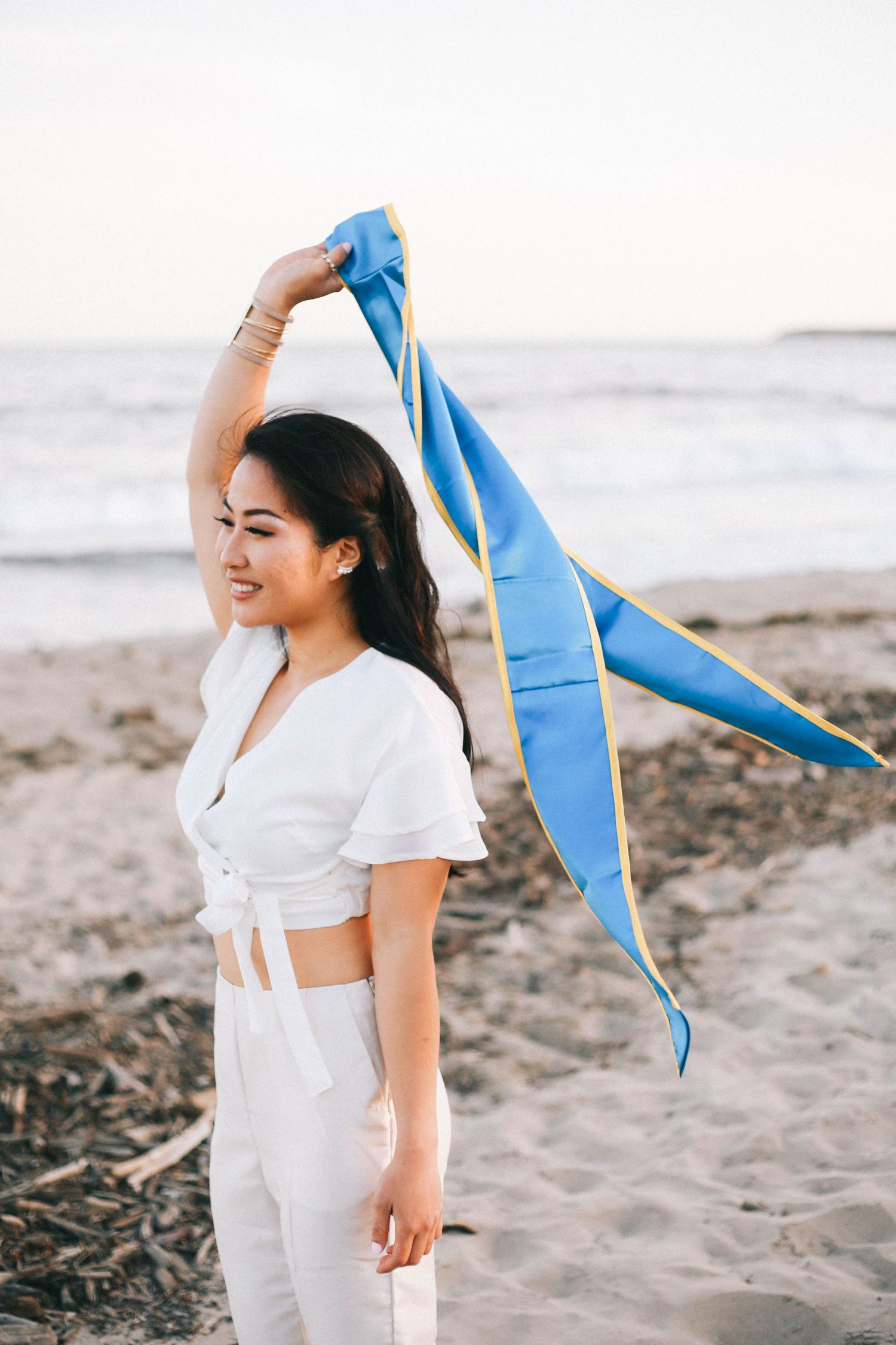 Young woman in white outfit holds blue and gold graduation stole on a sandy beach at sunset.