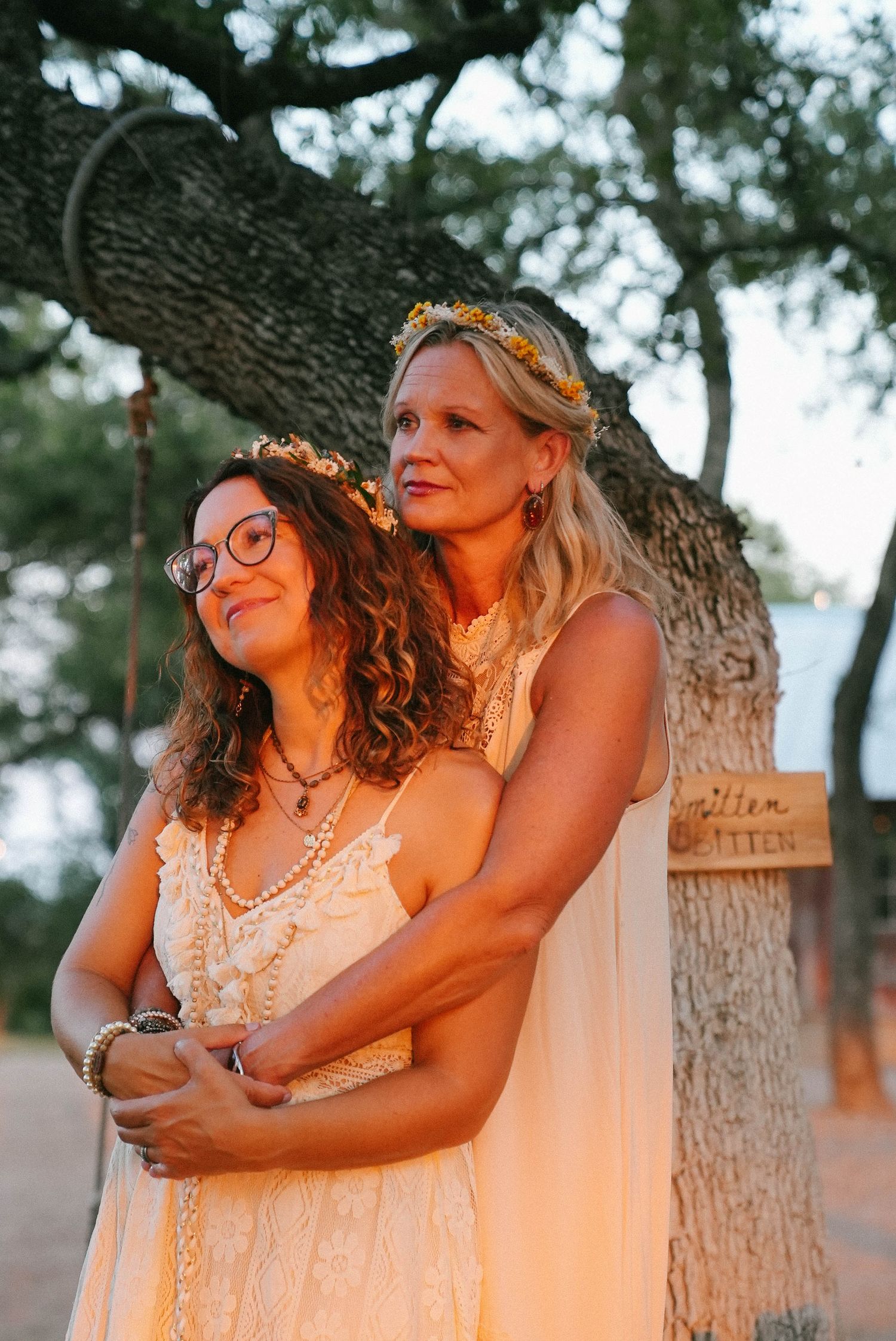 Two brides in white boho dresses with flower crowns embracing under oak tree at sunset.