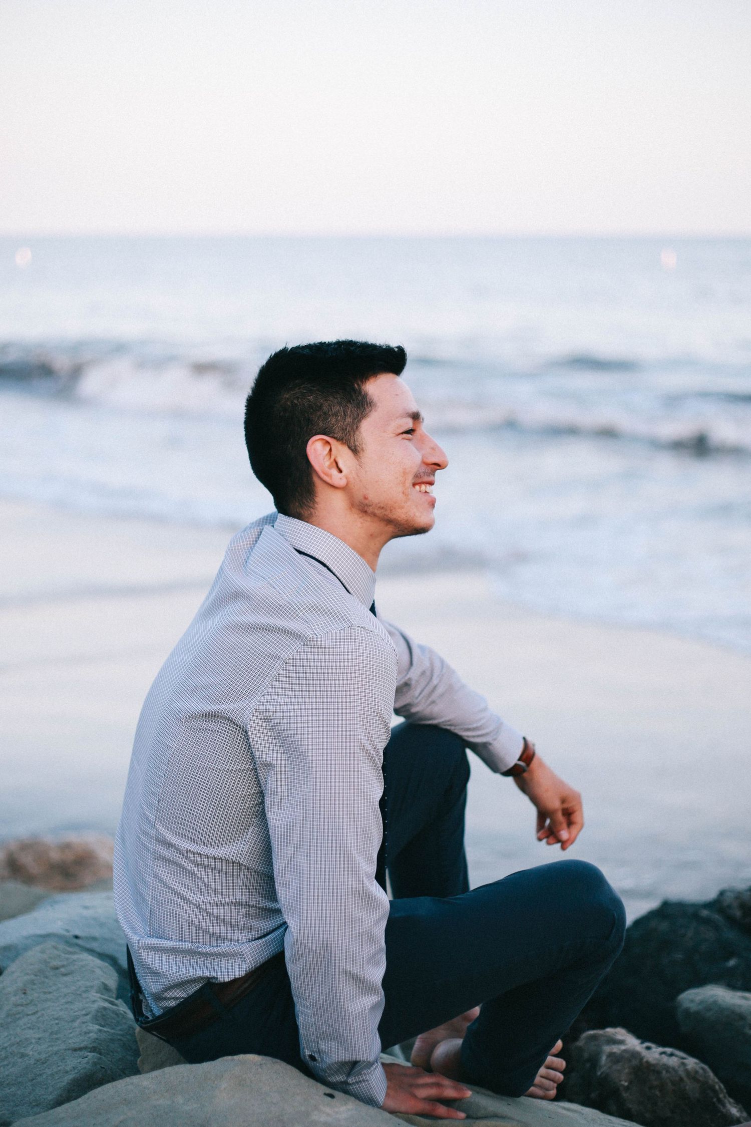 Young man in gray shirt sitting on rocks smiling at the ocean during golden hour.