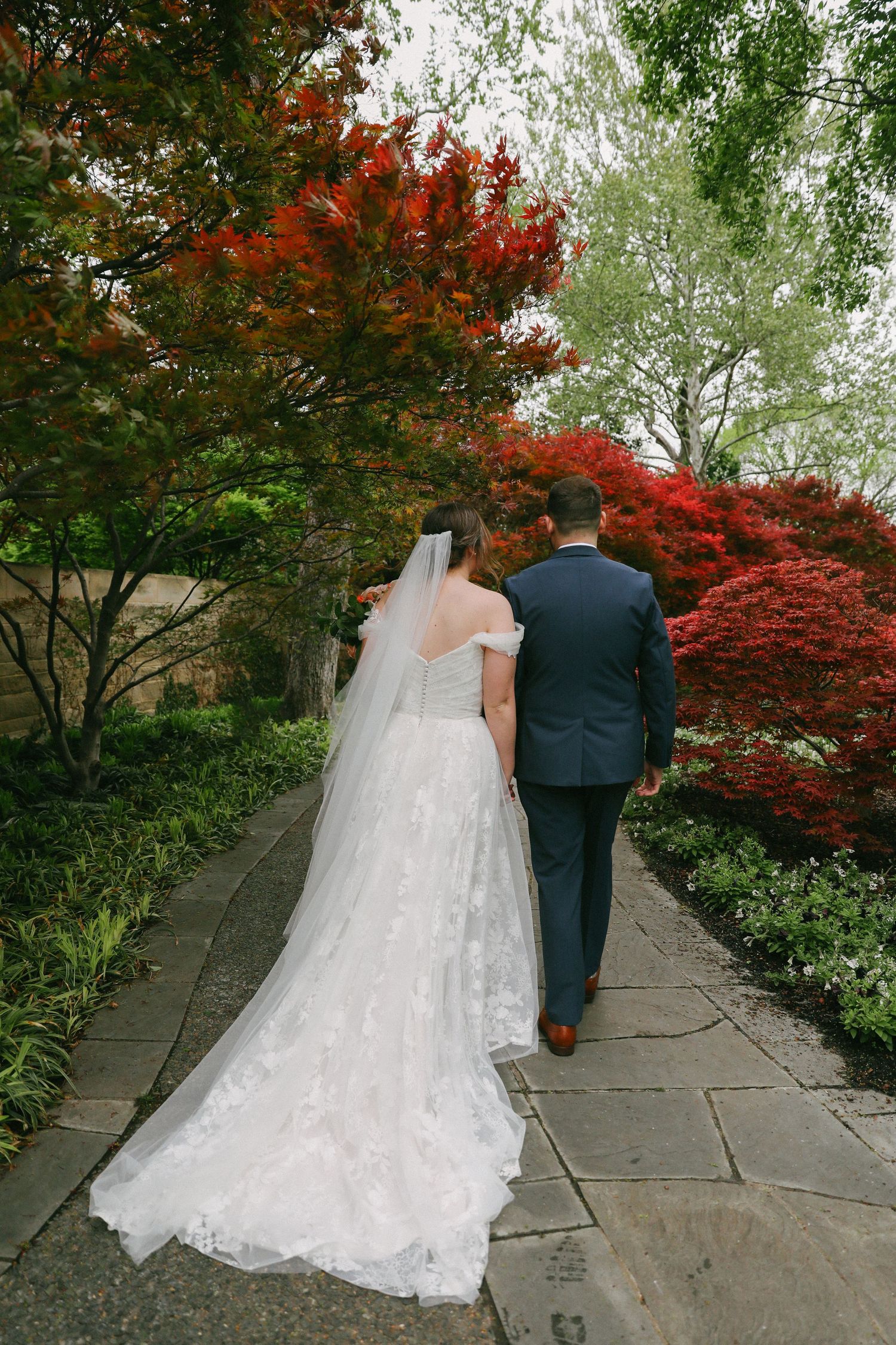 Bride and groom walk hand in hand down garden path lined with vibrant red Japanese maples.