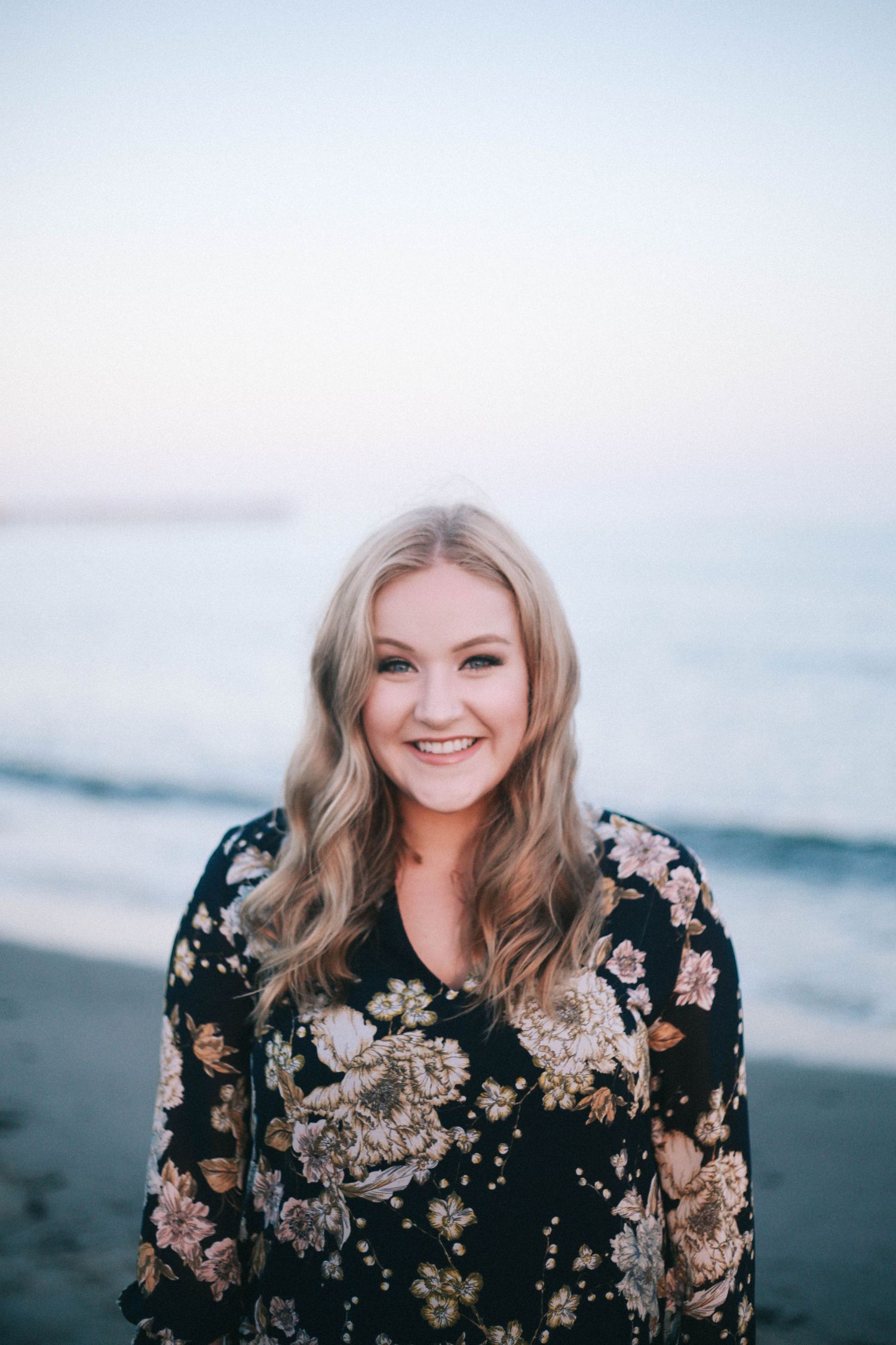 Smiling young blonde woman in a floral dress poses on a beach at dusk with ocean waves behind her.