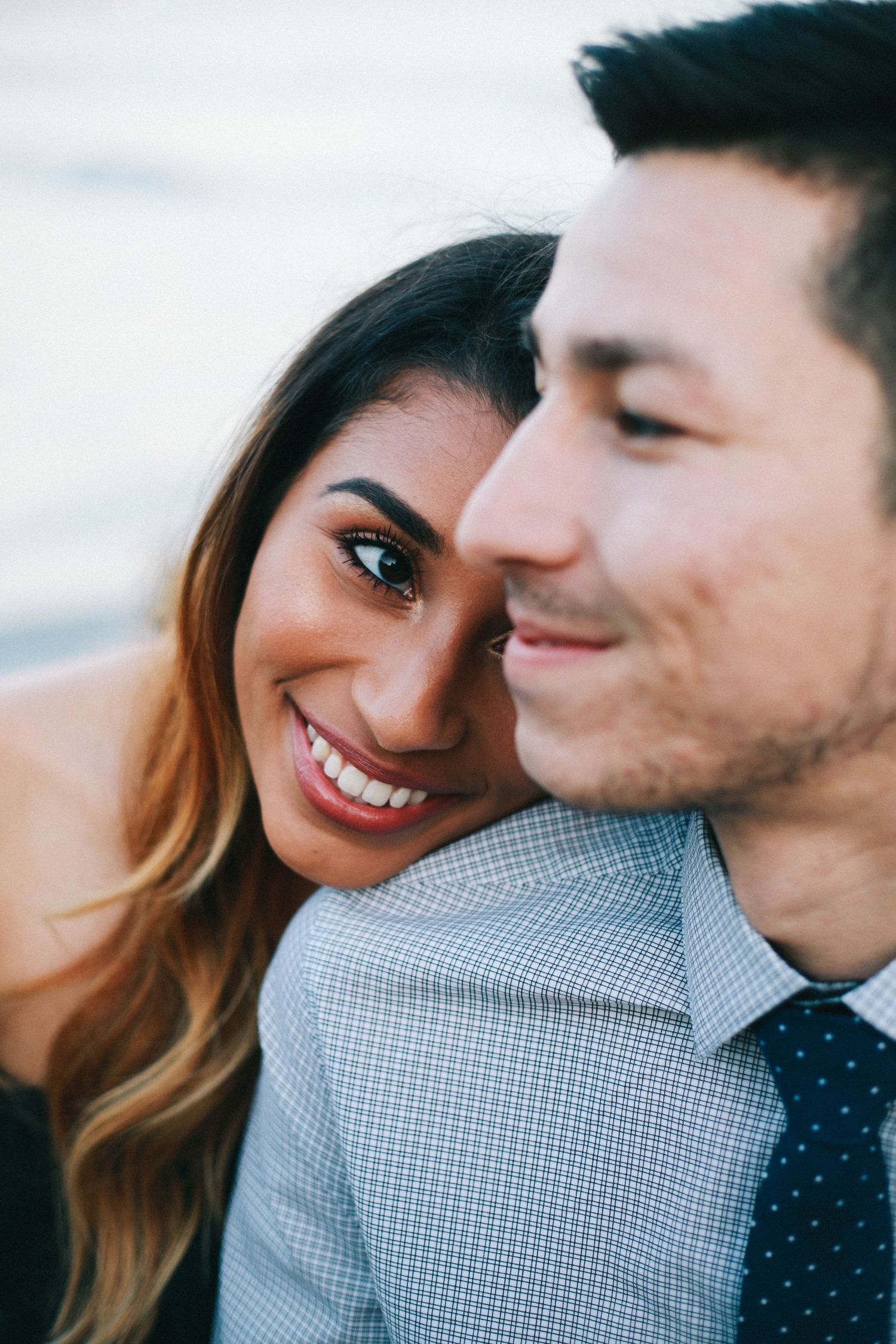 Close-up of smiling woman leaning on man in gray shirt and navy tie at the beach.