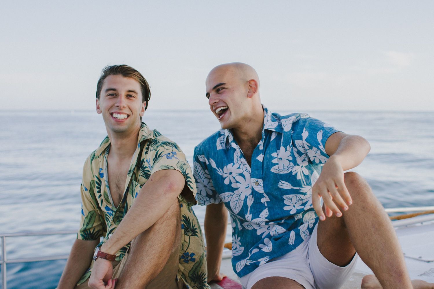 Two young men in Hawaiian shirts laughing together on a boat with the ocean behind them.