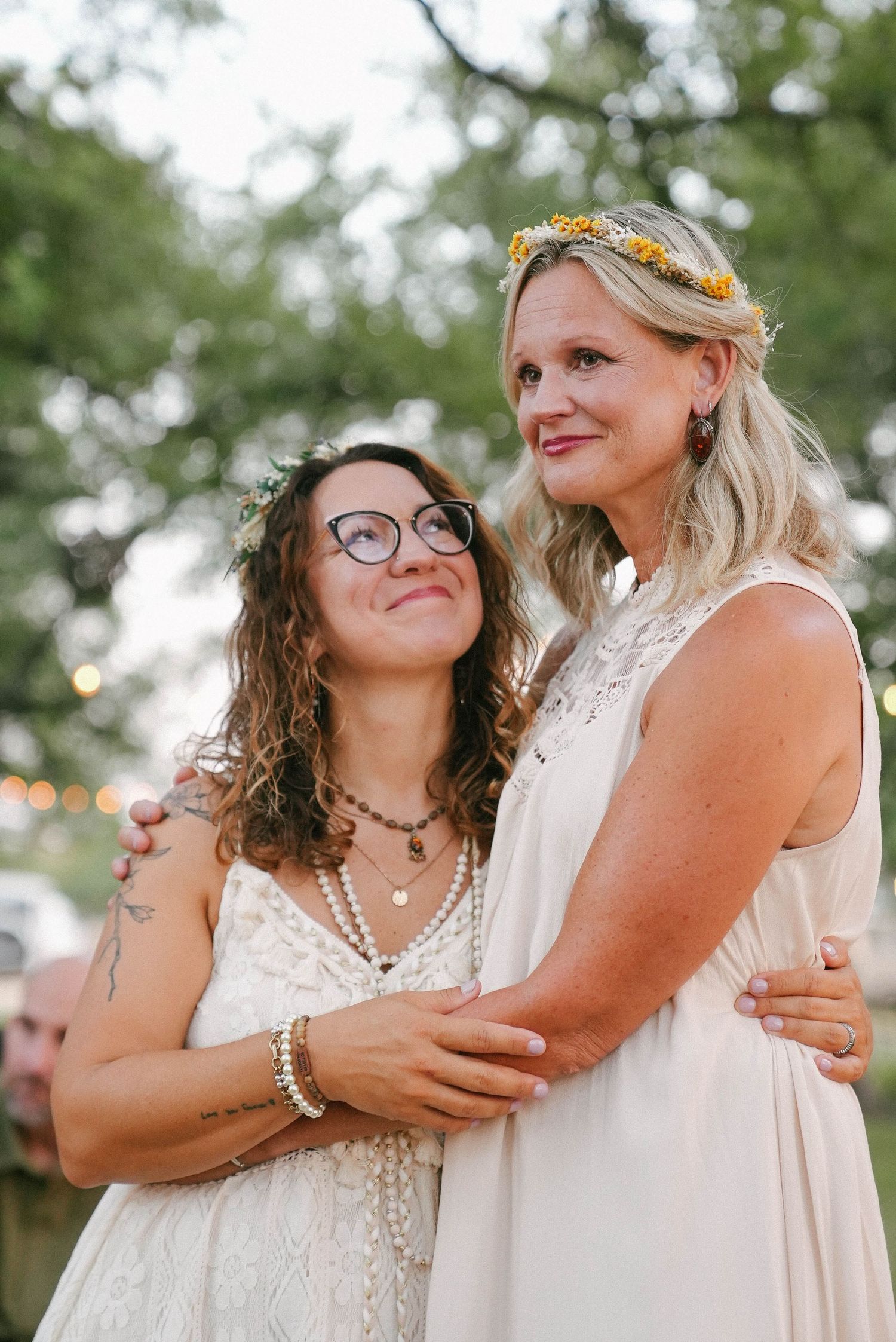 Two smiling brides in white boho dresses with flower crowns embracing at outdoor wedding.