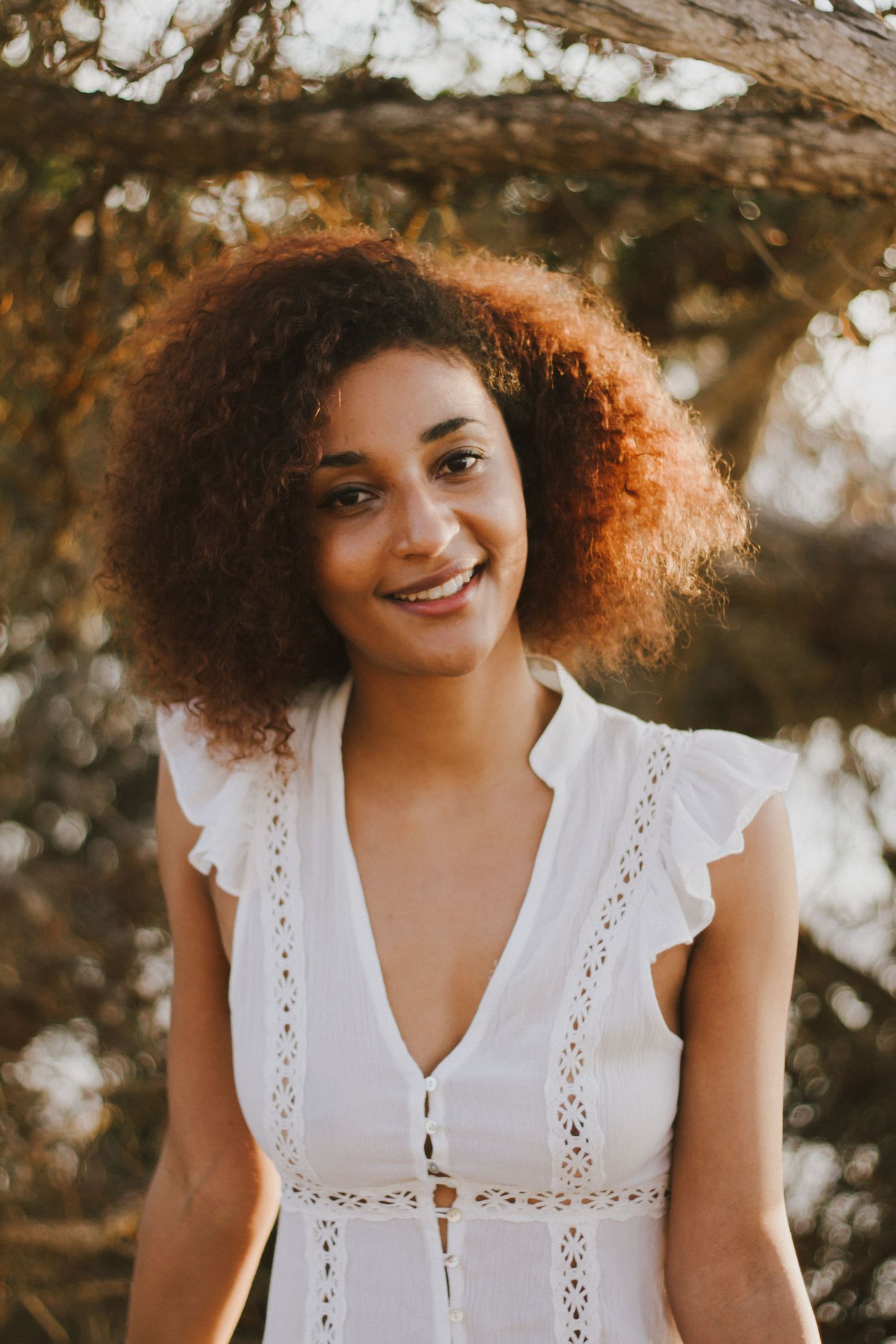 Young woman with curly auburn hair smiles in white dress under golden natural light outdoors.