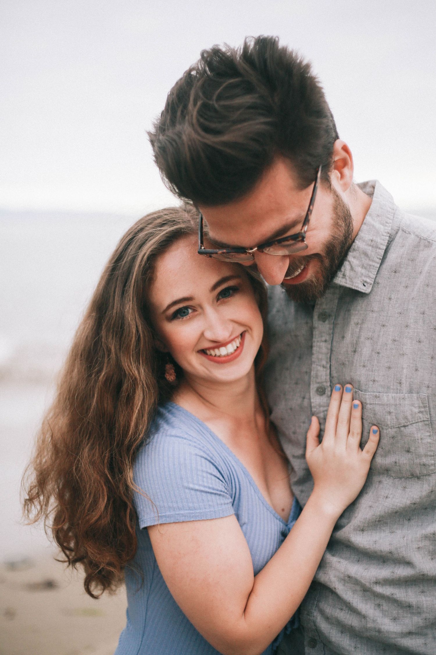 Smiling woman in blue top embraces bearded man with glasses near a beach.