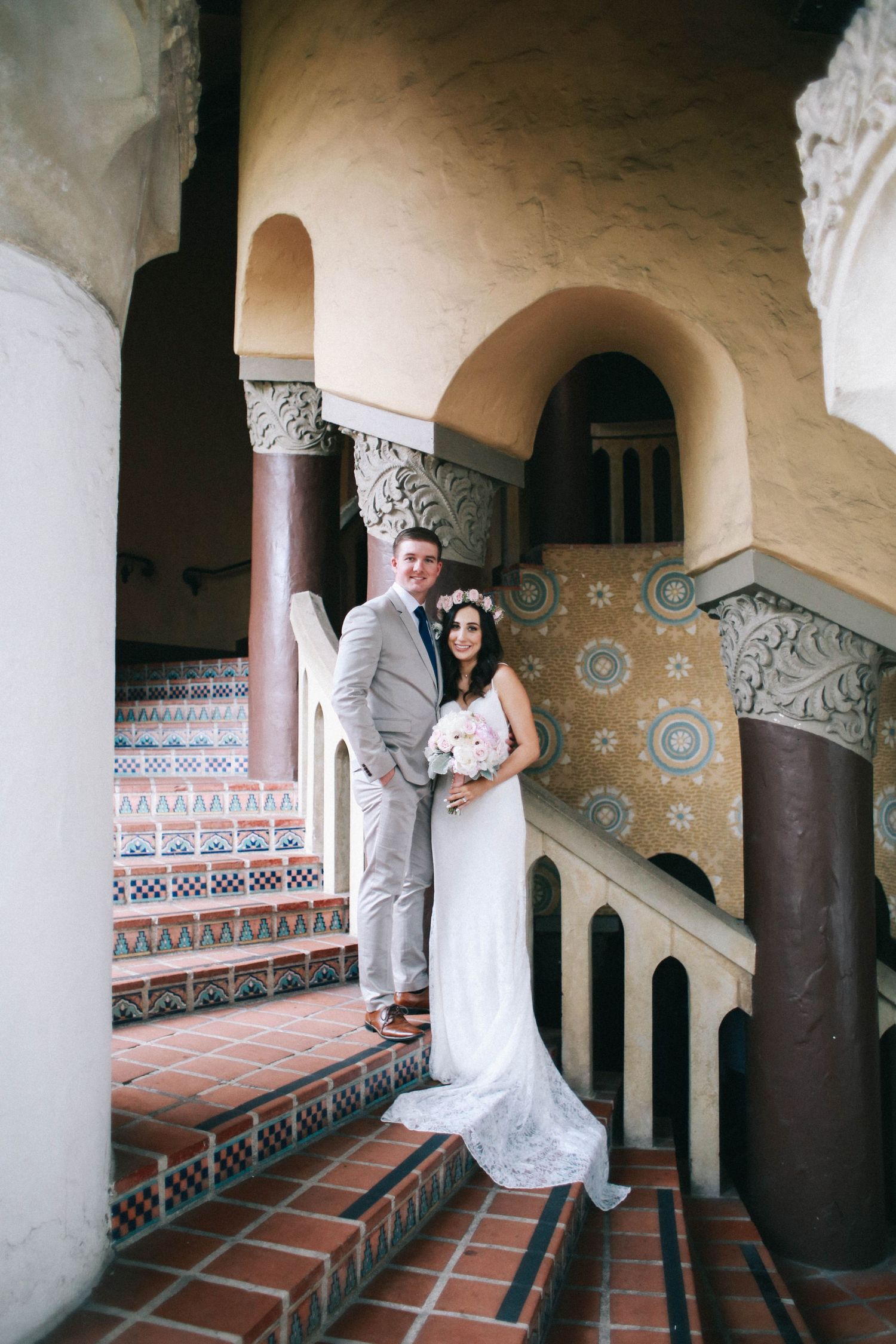 Bride and groom pose on ornate tiled staircase with arched columns at a Spanish-style wedding venue.