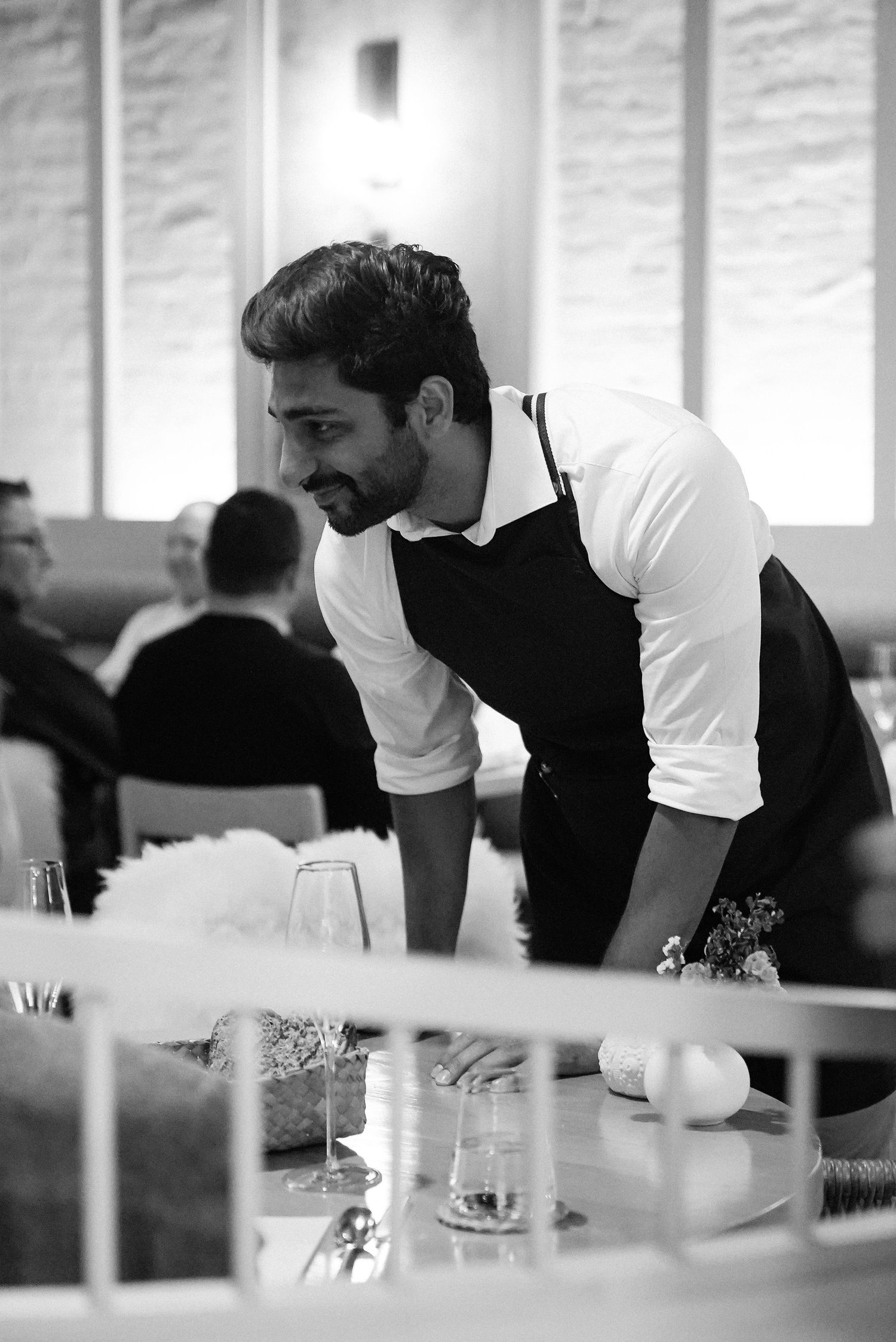 Attentive male server in black apron leans over table to assist diners in a modern restaurant.