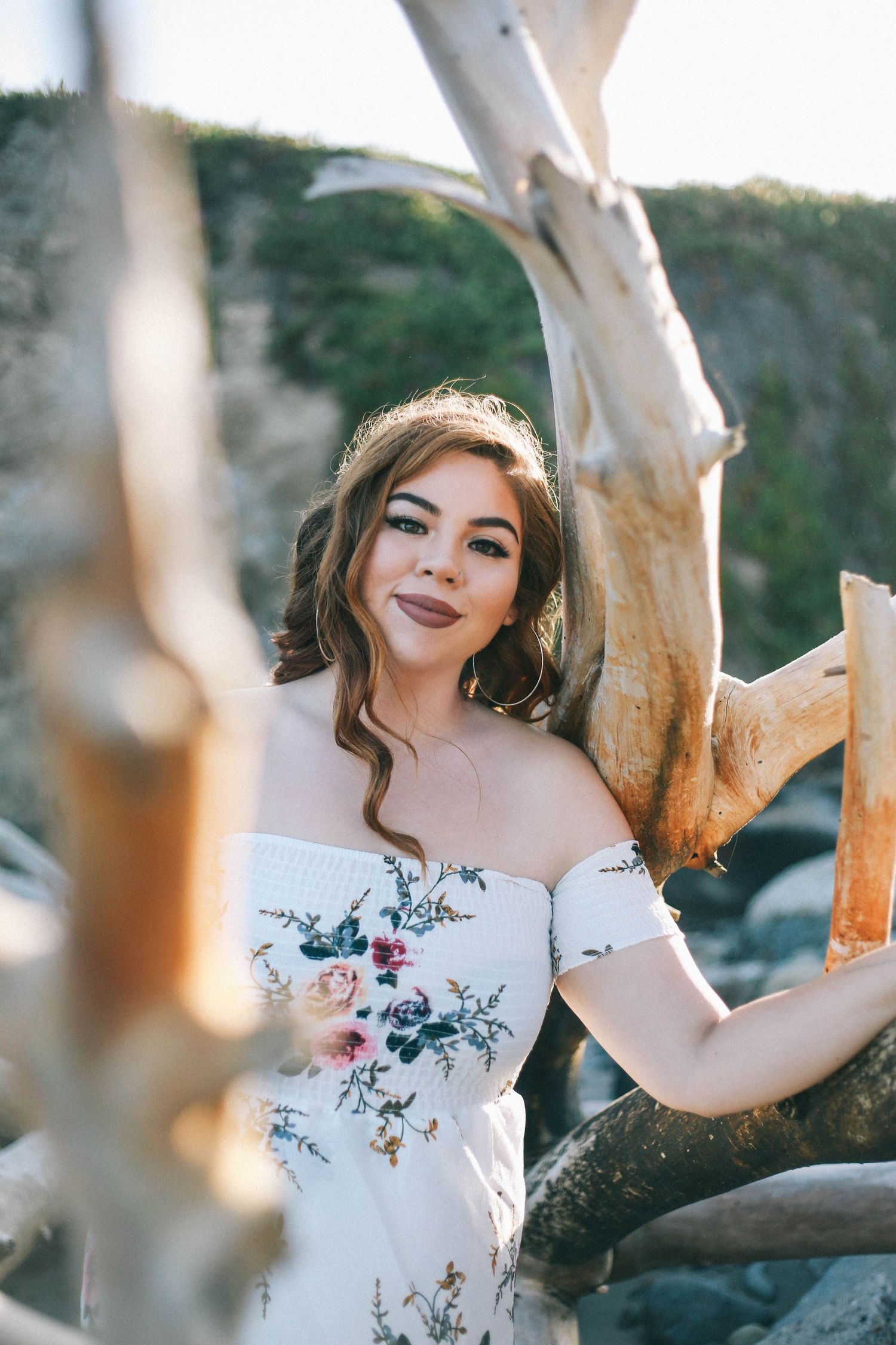 Young woman in off-shoulder floral dress posing near driftwood on a rocky beach.