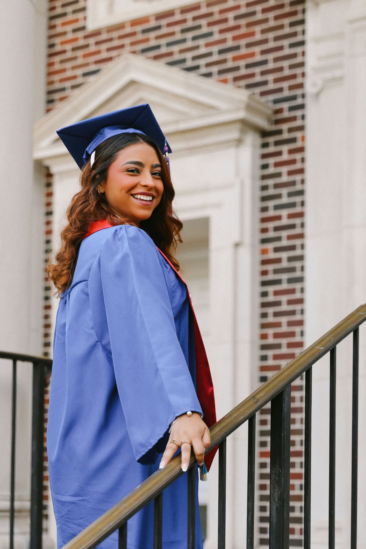 Smiling graduate in blue cap and gown on staircase railing in front of a brick building.