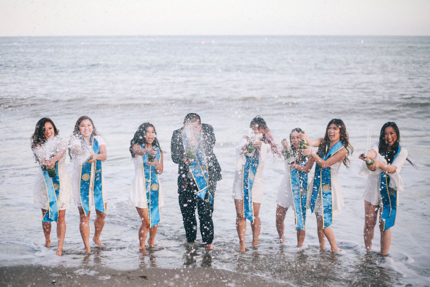 Group of graduates in blue sashes celebrating on the beach with confetti by the ocean.