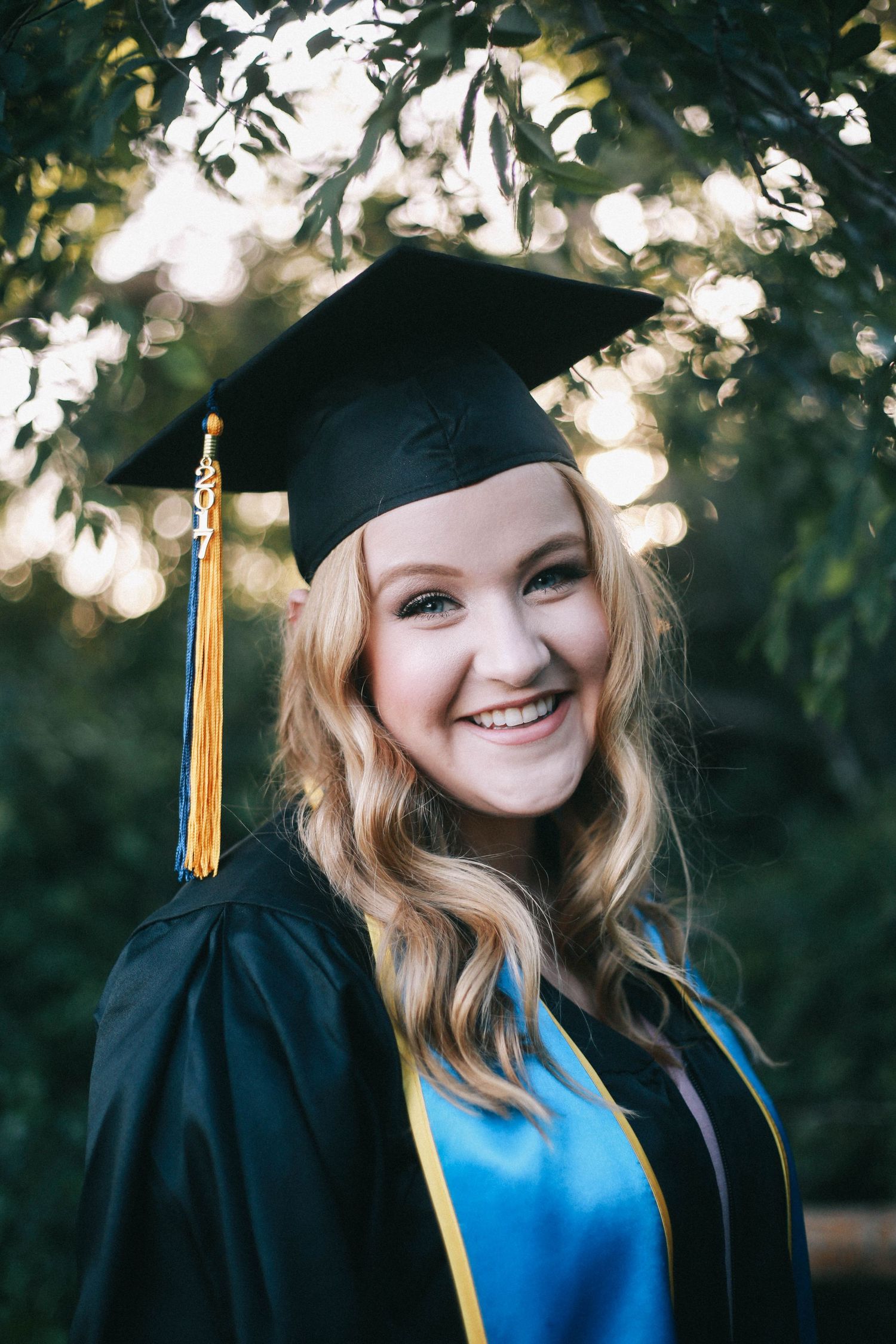 Happy blonde graduate smiling in black cap and gown with blue and gold accents outdoors.