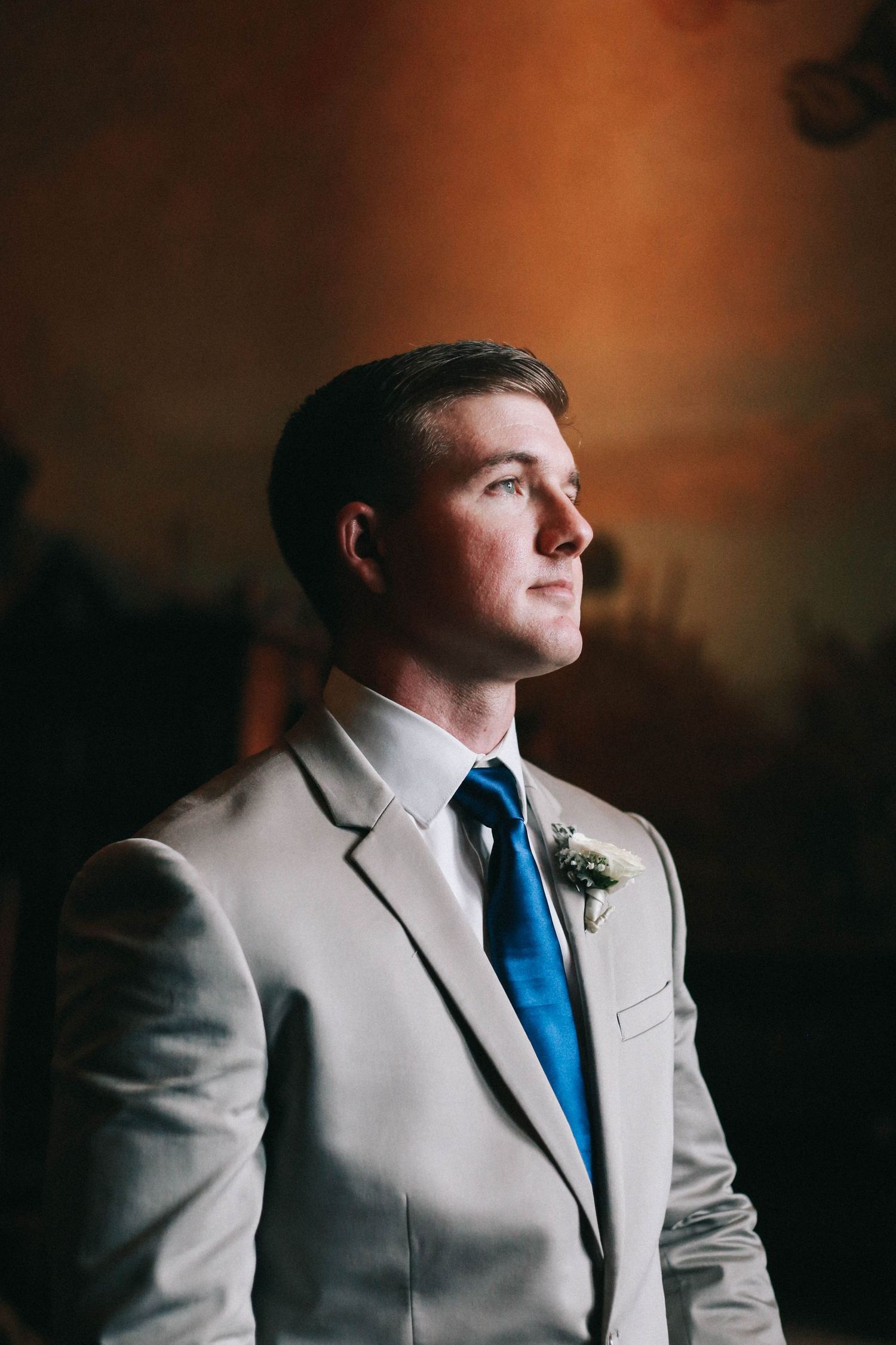 Elegant groom in gray suit and blue tie gazing upward in dramatic warm lighting.