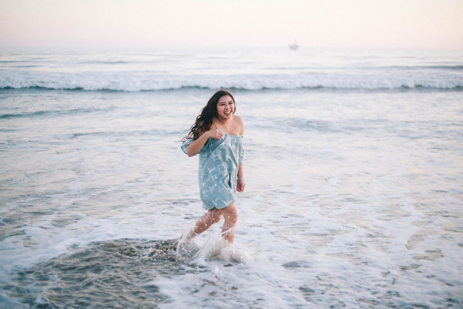 Happy young woman in blue dress splashing in ocean waves at sunset beach.