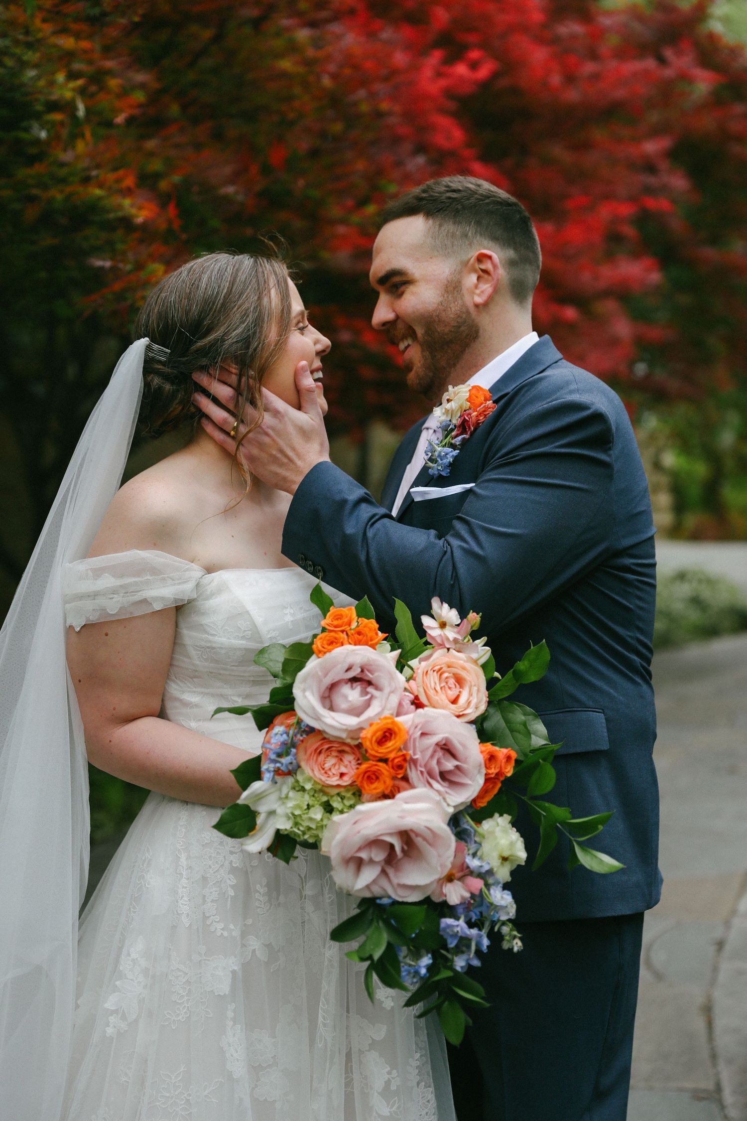 Happy bride and groom share a romantic moment with a colorful bouquet of orange and pink roses at an outdoor wedding.