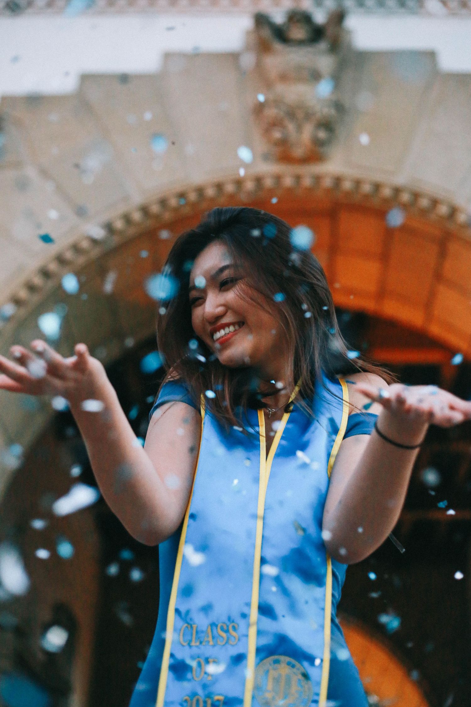 Joyful graduate in blue cap and gown tosses confetti outside an ornate university building.