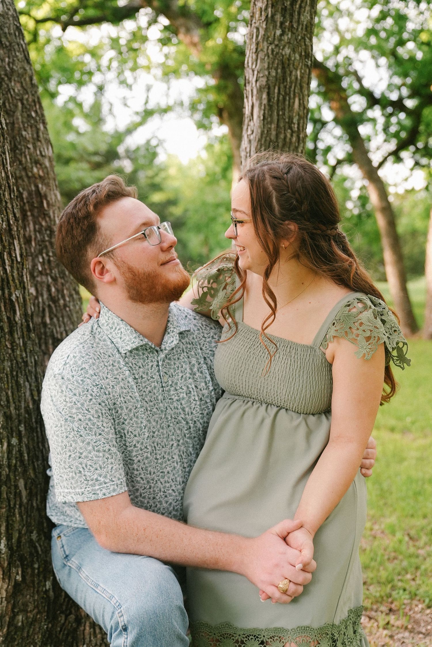 Couple sitting together against tree trunk, holding hands and smiling at each other outdoors.
