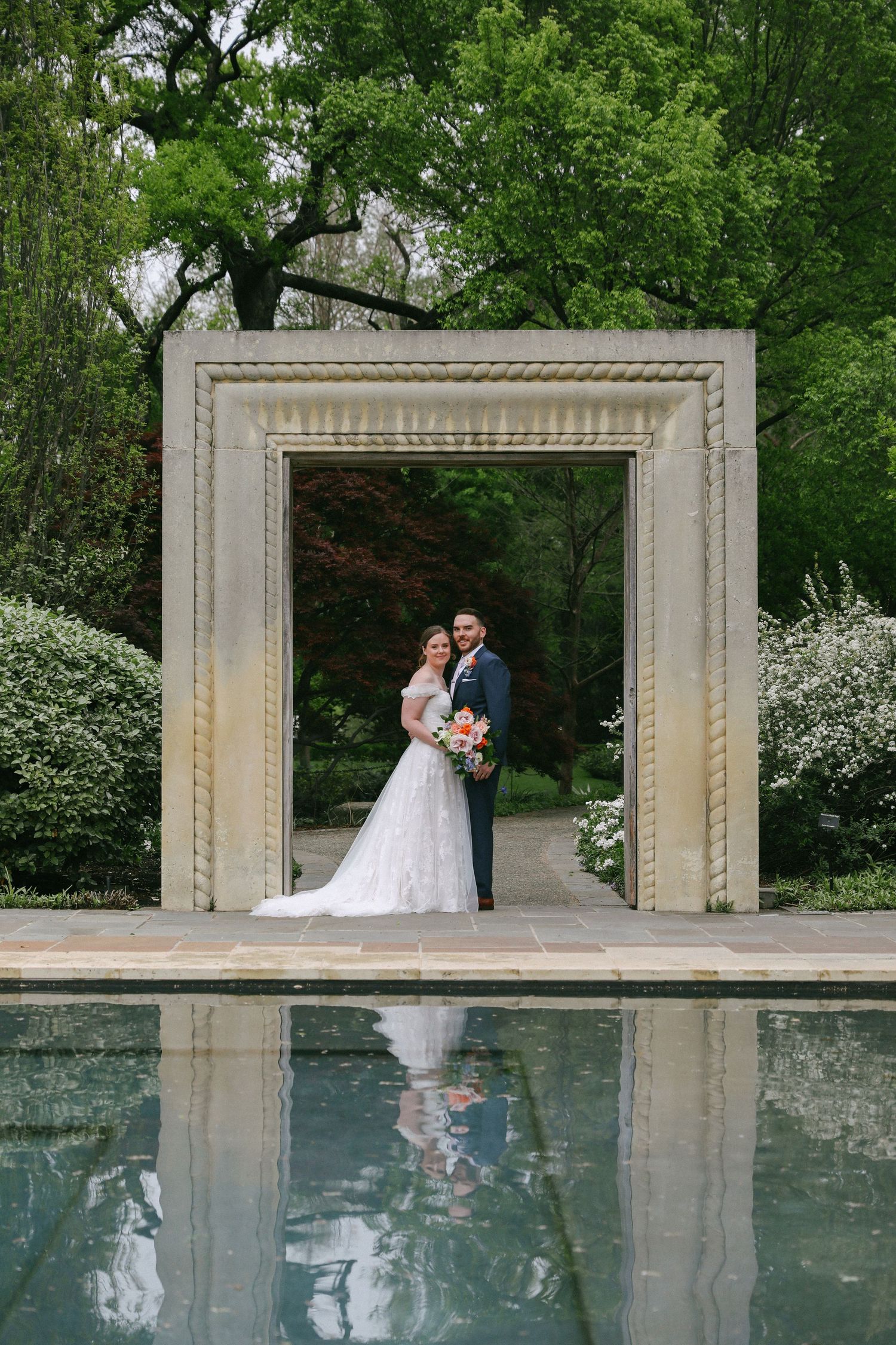 Bride and groom pose under stone arch reflected in still pool surrounded by lush gardens.