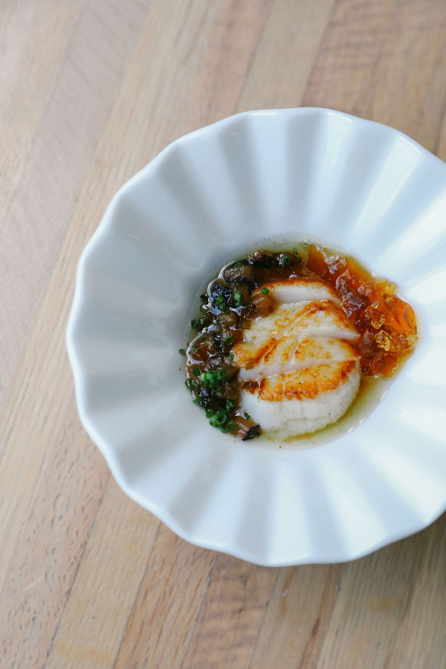 Seared scallop with capers, citrus sauce and herbs in white scalloped fine dining bowl.