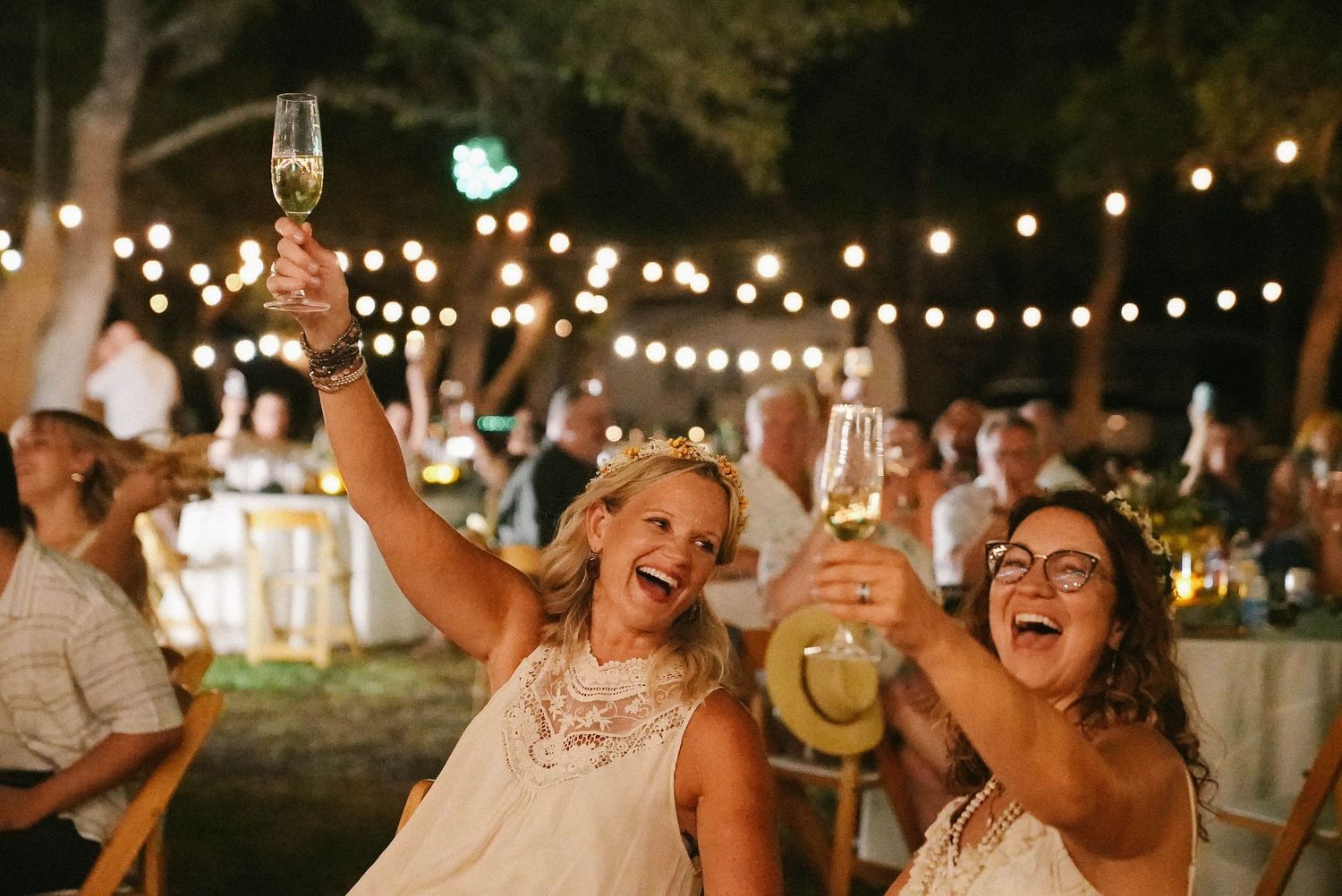 Two women in white dresses joyfully raise champagne glasses at an outdoor wedding reception with string lights.
