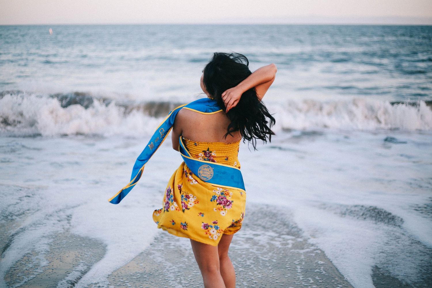 Woman in yellow floral dress with blue graduation sash standing at the ocean's edge.