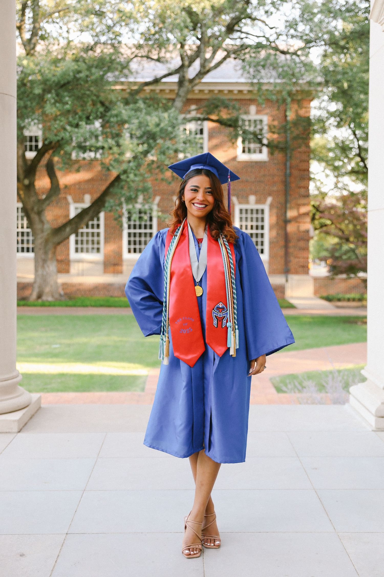 Smiling graduate in blue cap and gown with red stole and medals standing on a campus walkway.