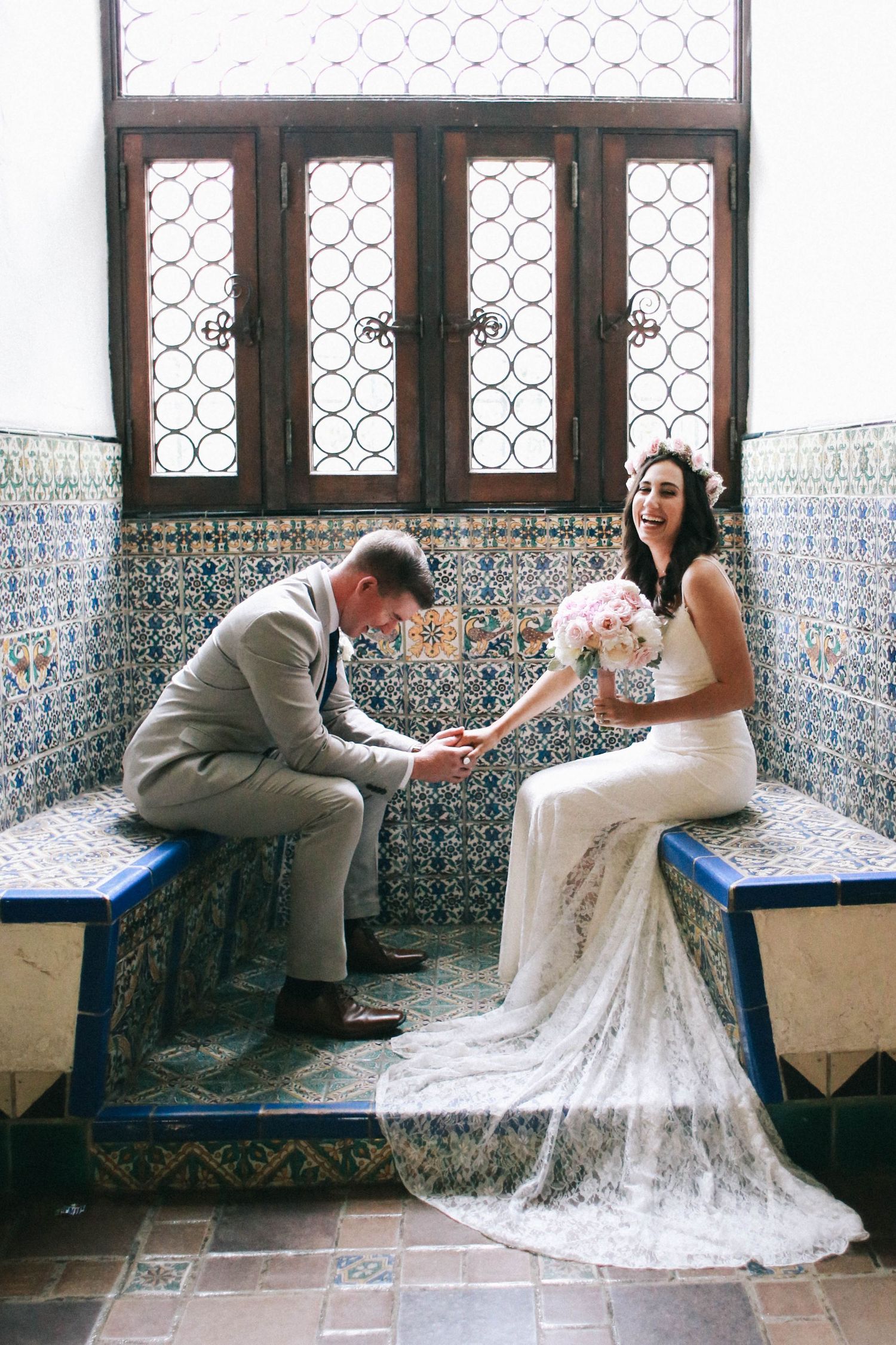 Bride and groom sitting on ornate tiled bench, sharing a romantic moment with pink bouquet indoors.