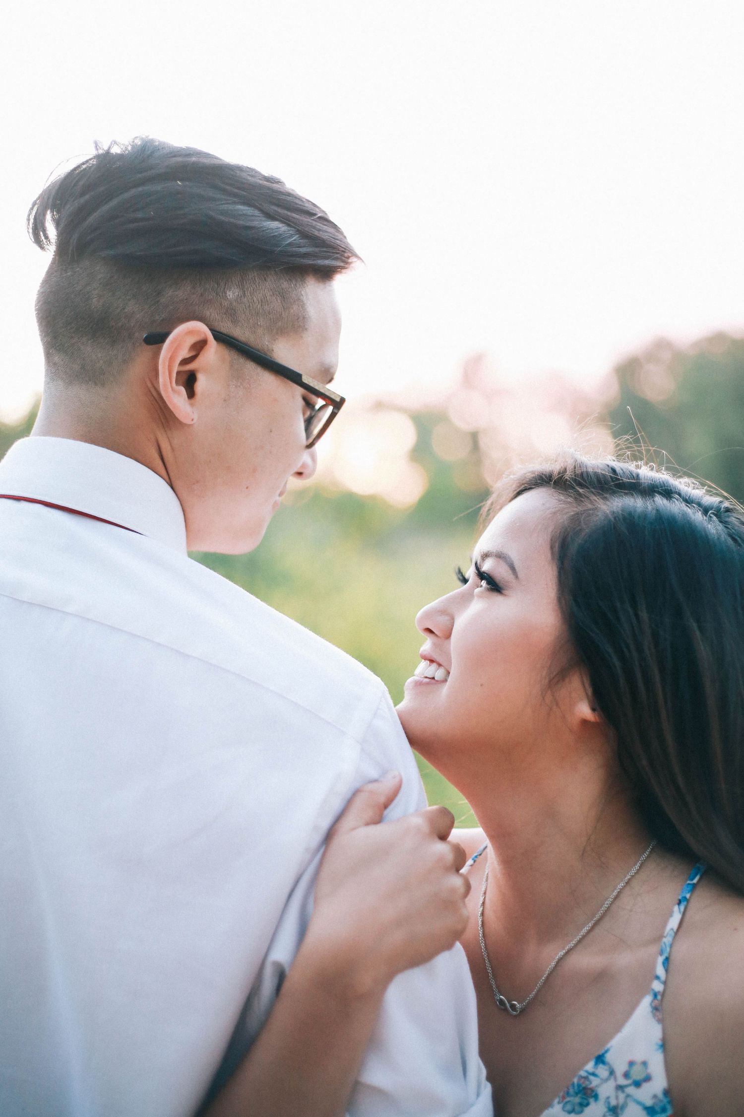 A smiling young woman gazes up at her partner while holding his shirt in a sunlit outdoor setting.