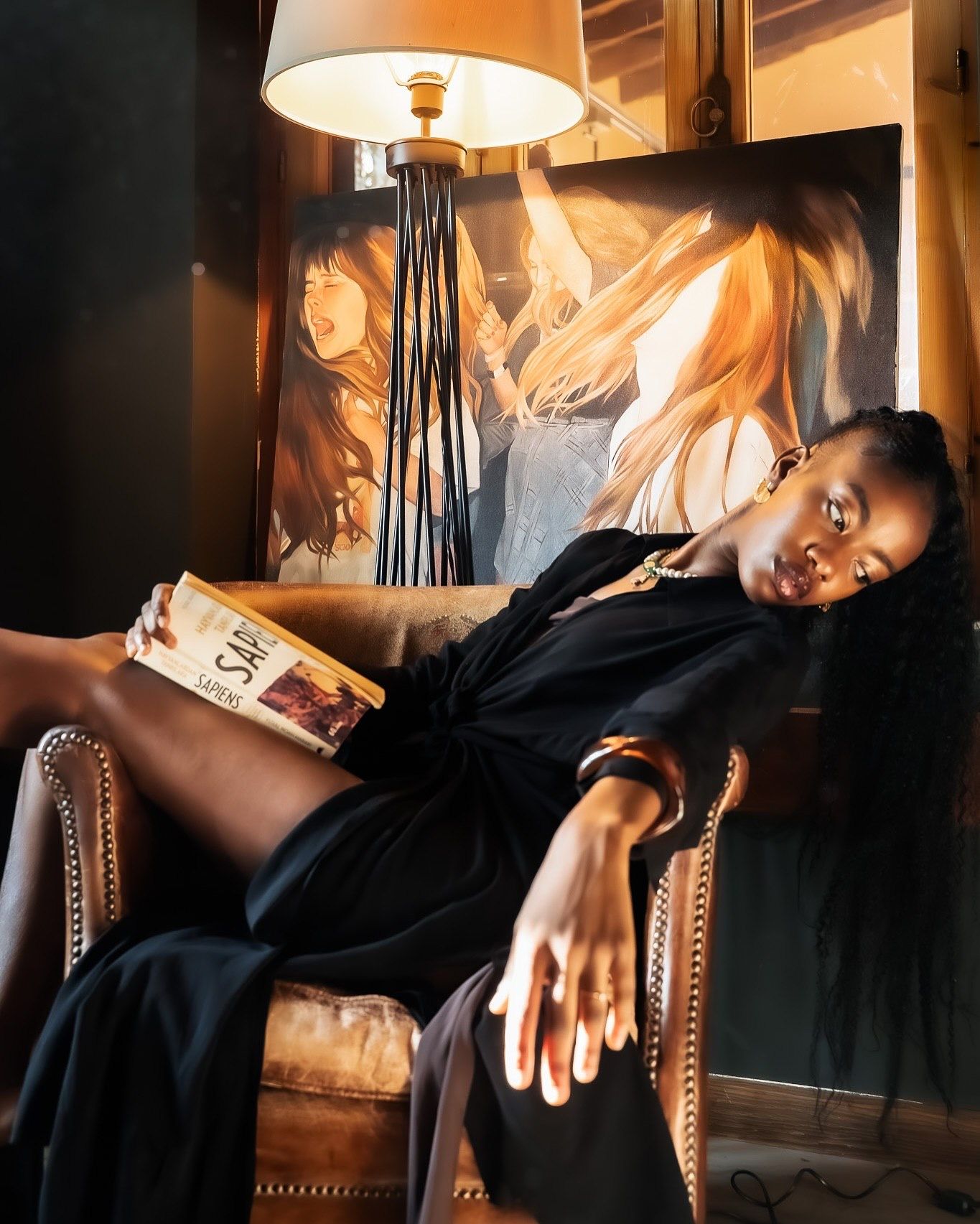 A woman in black dress reclines in leather chair holding book, beside lamp and painting in warm light.