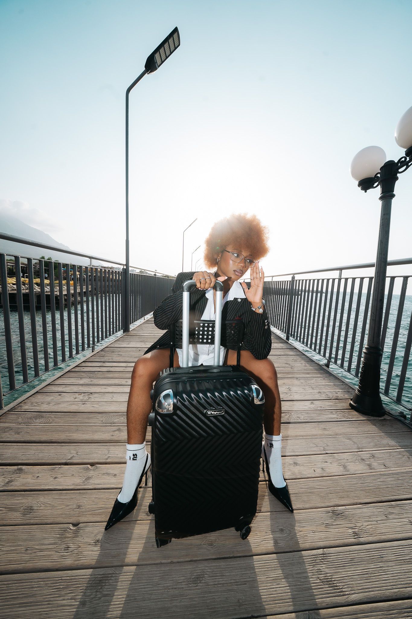Fashionable woman with afro sits on black suitcase on wooden pier wearing pinstripe blazer.