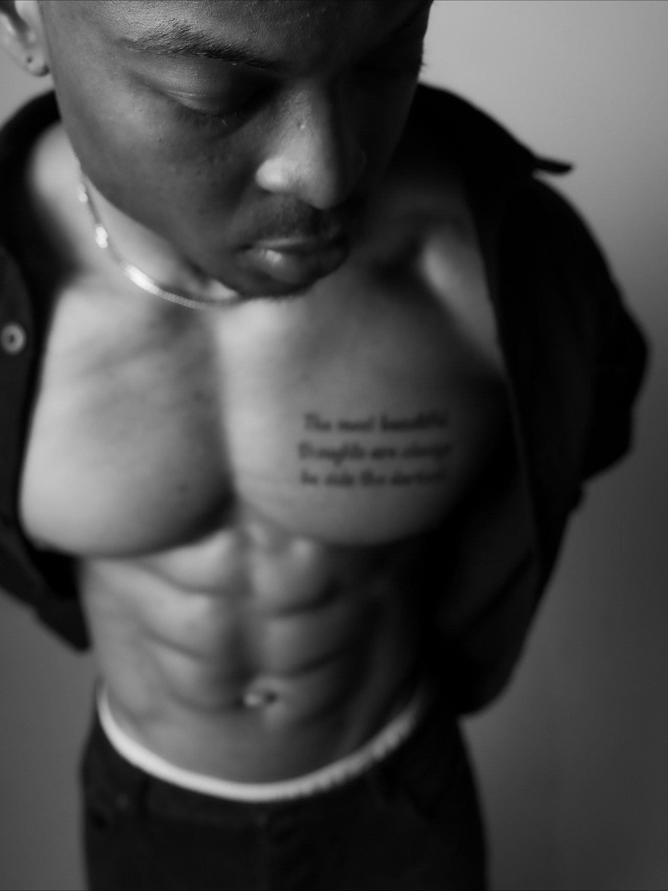 Black and white photo of a shirtless muscular man with a chest tattoo, wearing an open black shirt and silver necklace.