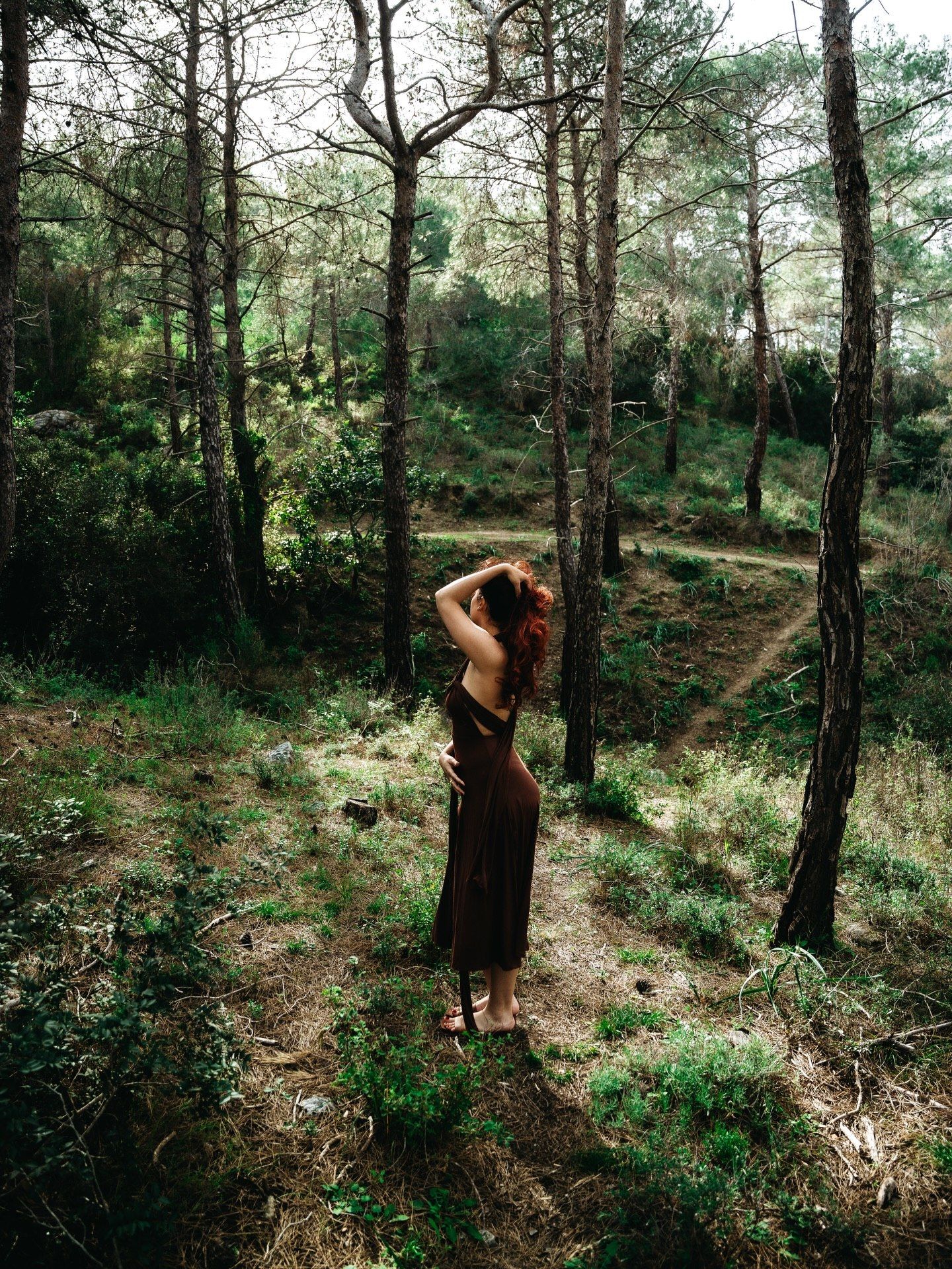 Woman in dark dress stands barefoot among tall trees in a sunlit forest clearing.