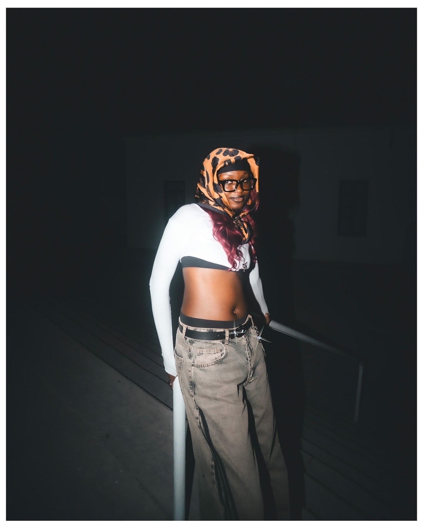Person wearing leopard print headscarf, glasses, white crop top, and baggy jeans posing in dark setting.