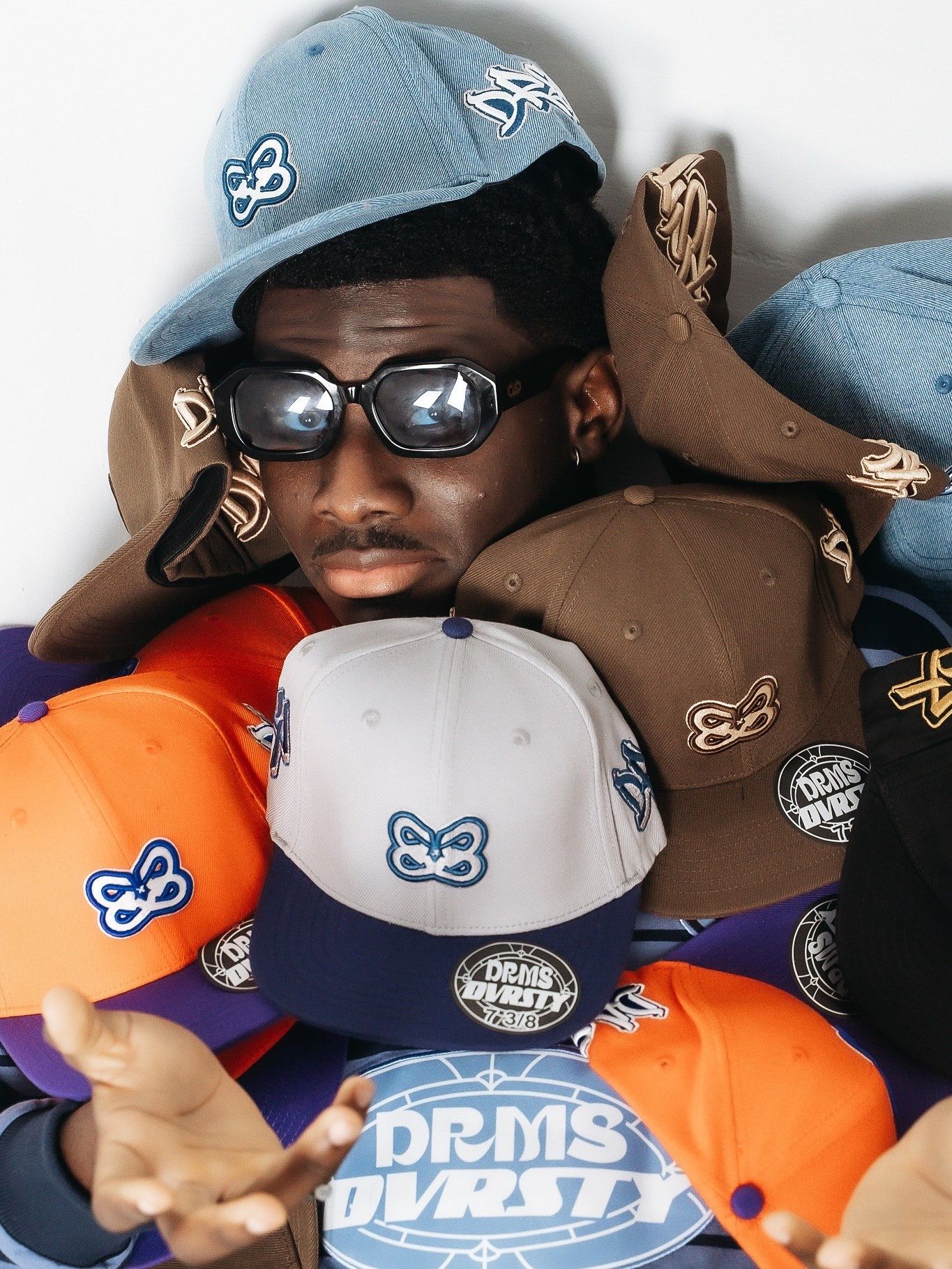 Person surrounded by colorful snapback caps featuring butterfly logos and DRMS DVRST branding in blue, orange, brown, and gray.