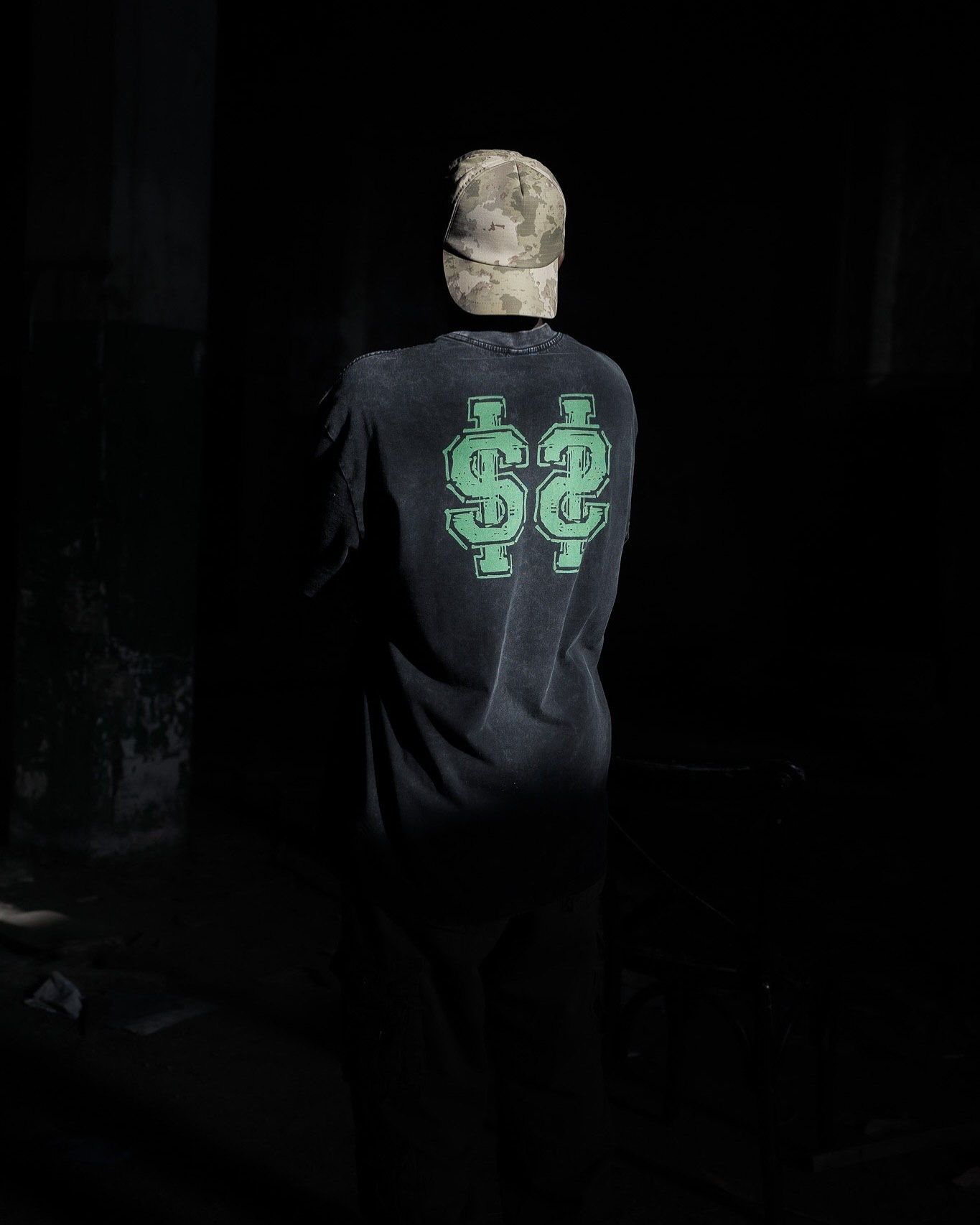 Person wearing black long-sleeve shirt with green dollar sign logo on back, standing in dark setting with camo cap.