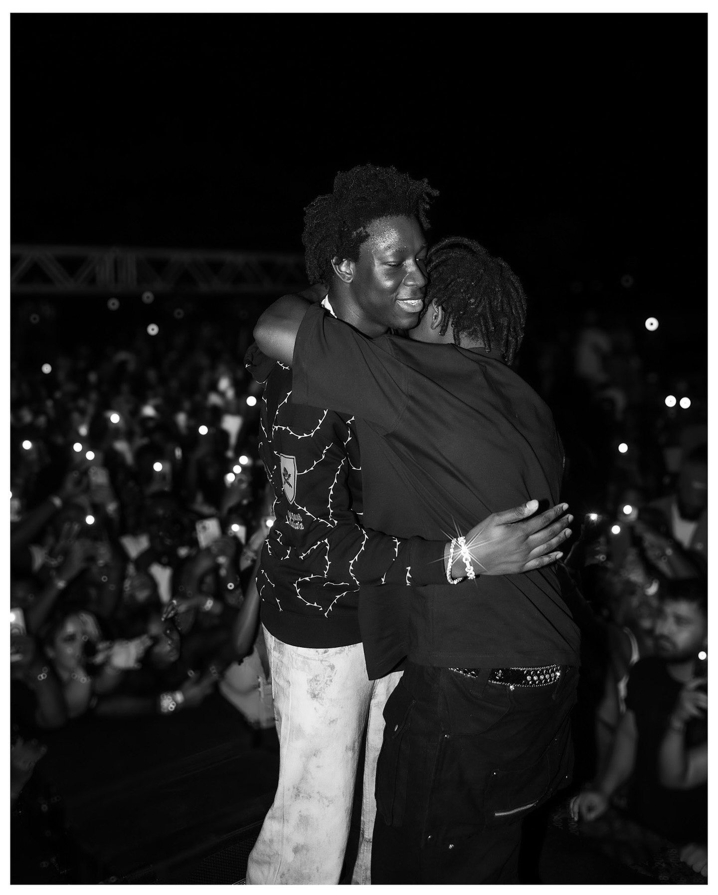 Two performers share an emotional hug on stage at a night concert, with a crowd of fans holding up phone lights in the background.