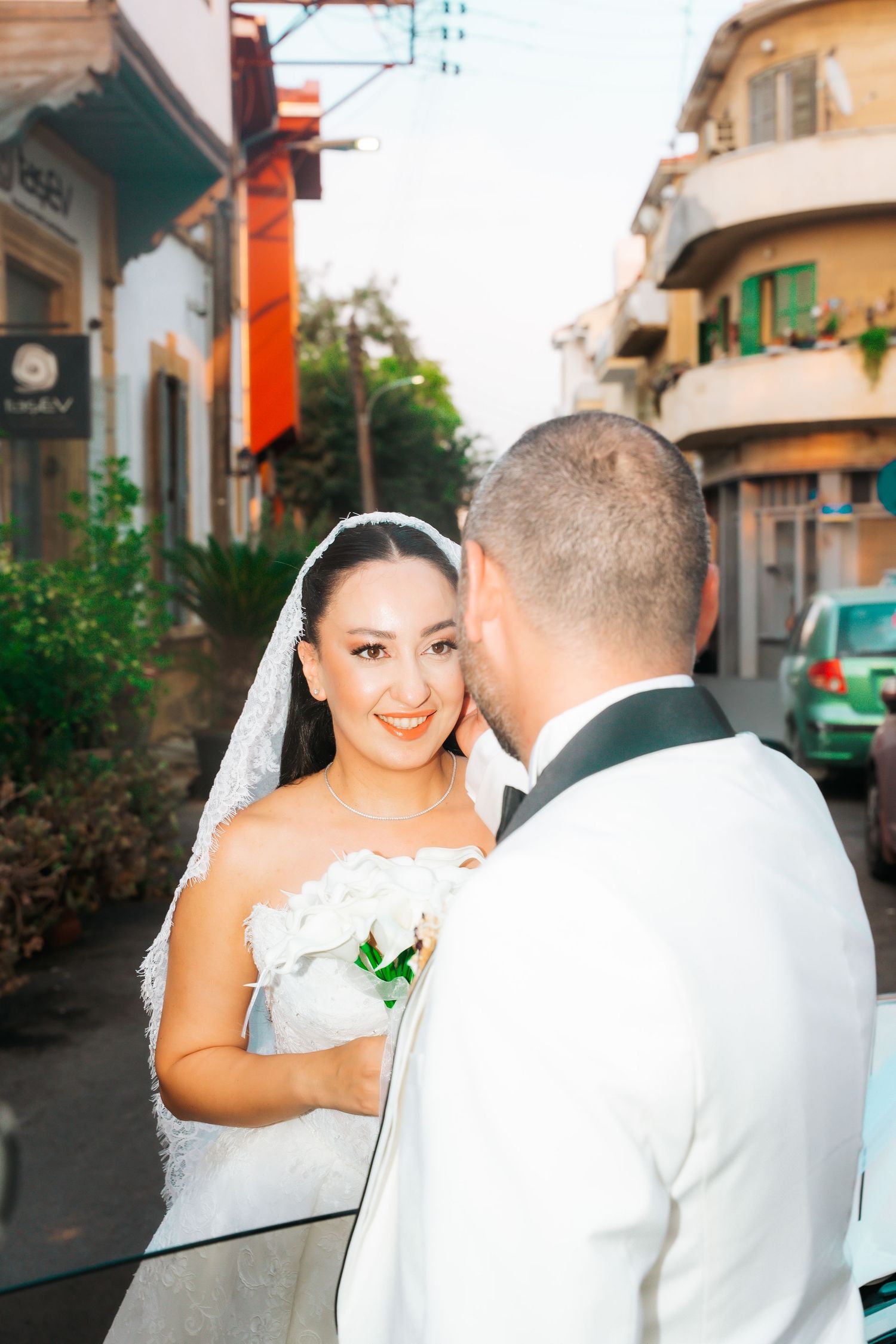 Smiling bride in white gown and veil holds bouquet facing groom in white tuxedo on a charming street.