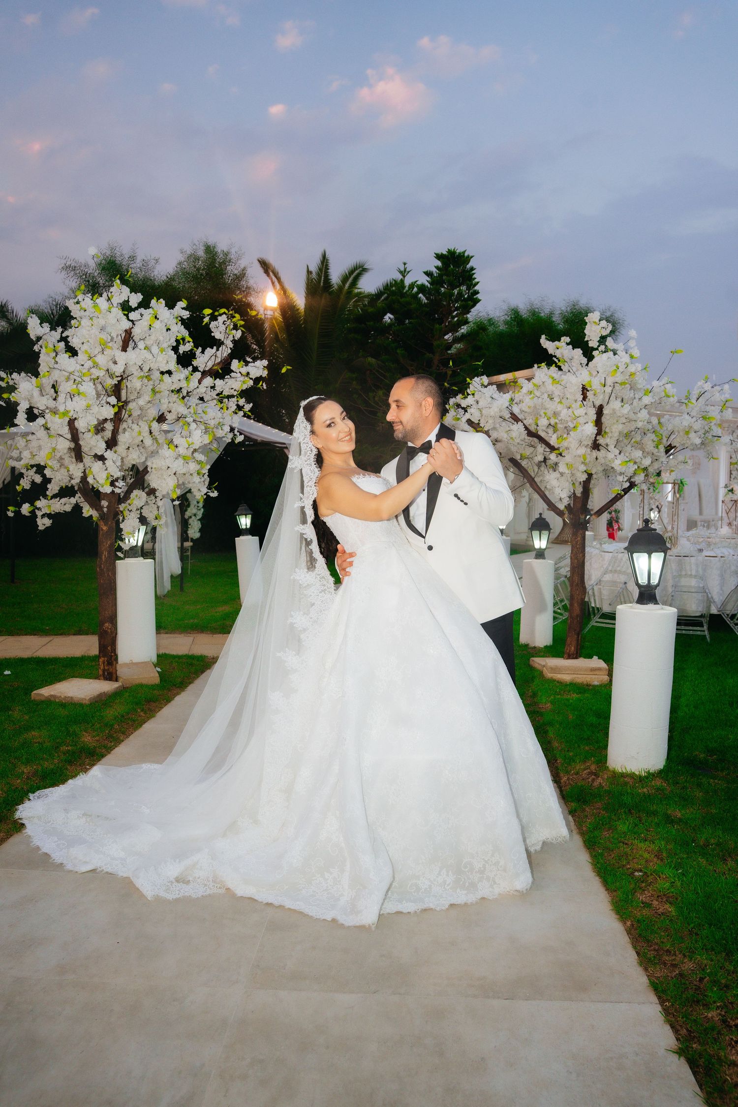 Bride and groom embrace outdoors at dusk, surrounded by white blossom trees and lanterns at elegant wedding venue.