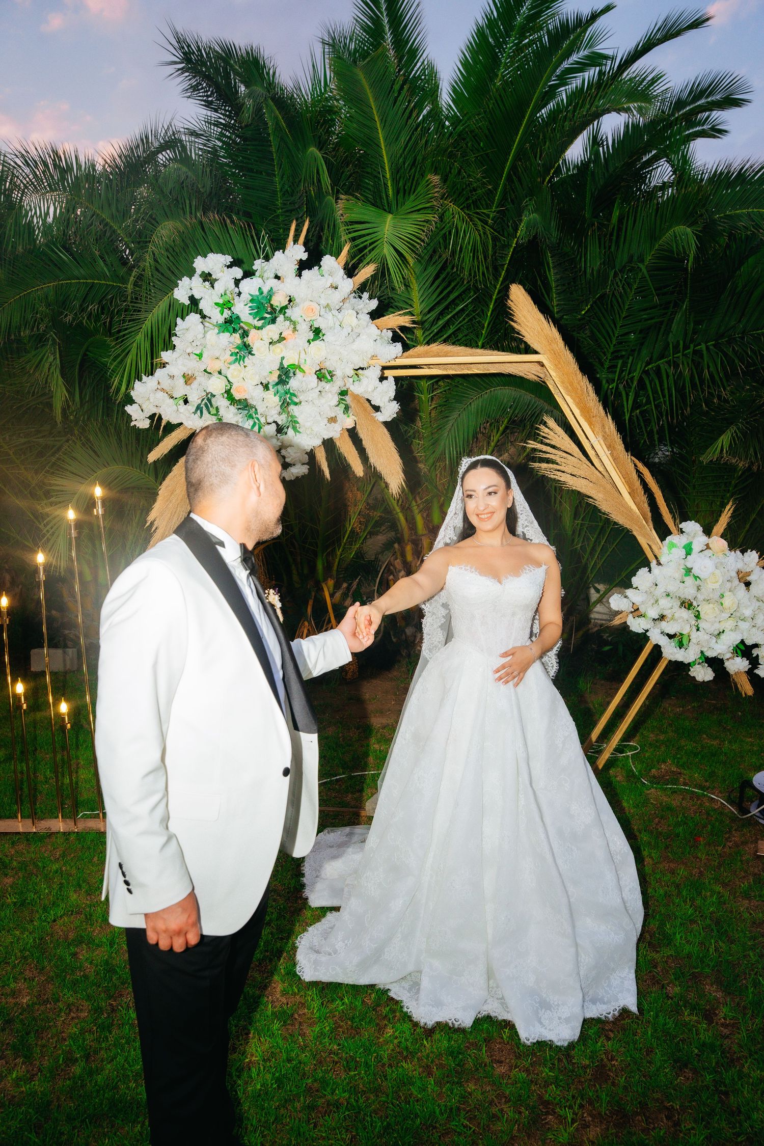 Bride and groom holding hands at outdoor wedding ceremony under floral arch with white flowers and palm trees at dusk.