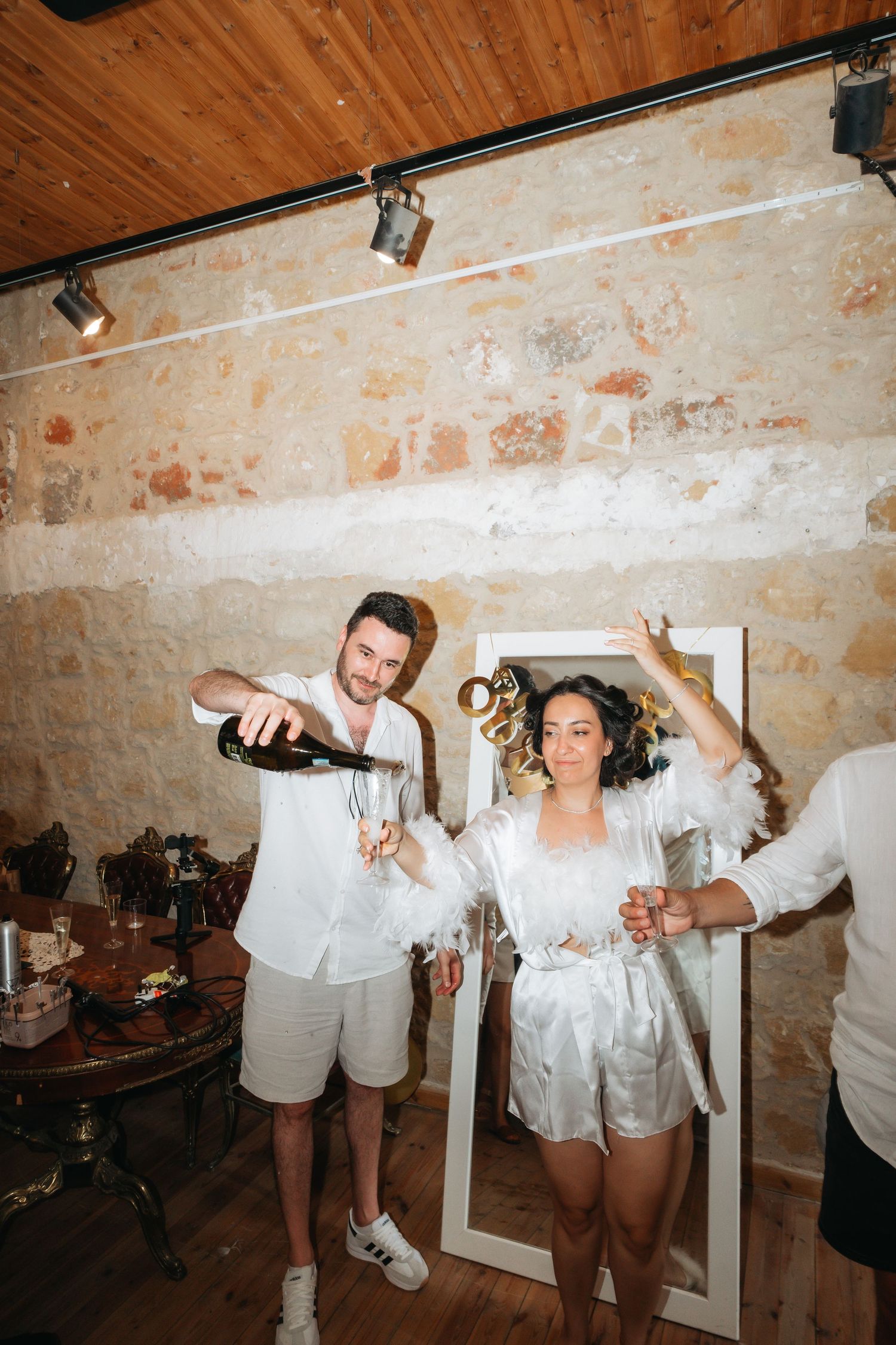 Couple celebrating with champagne in a rustic stone-walled room, woman in white robe holding glass up joyfully.