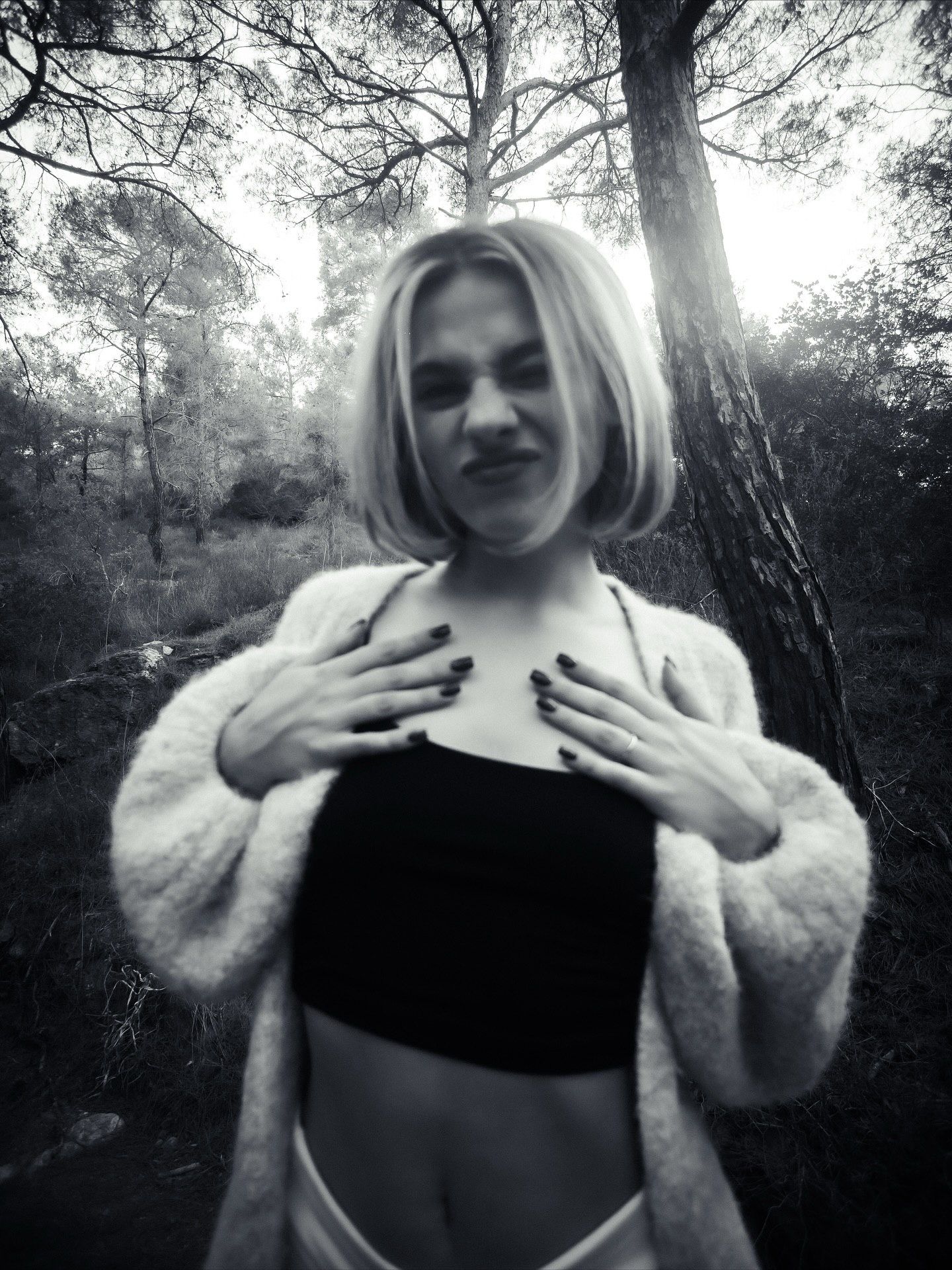 Black and white photo of a blonde woman in a fur coat and crop top posing in a forest.