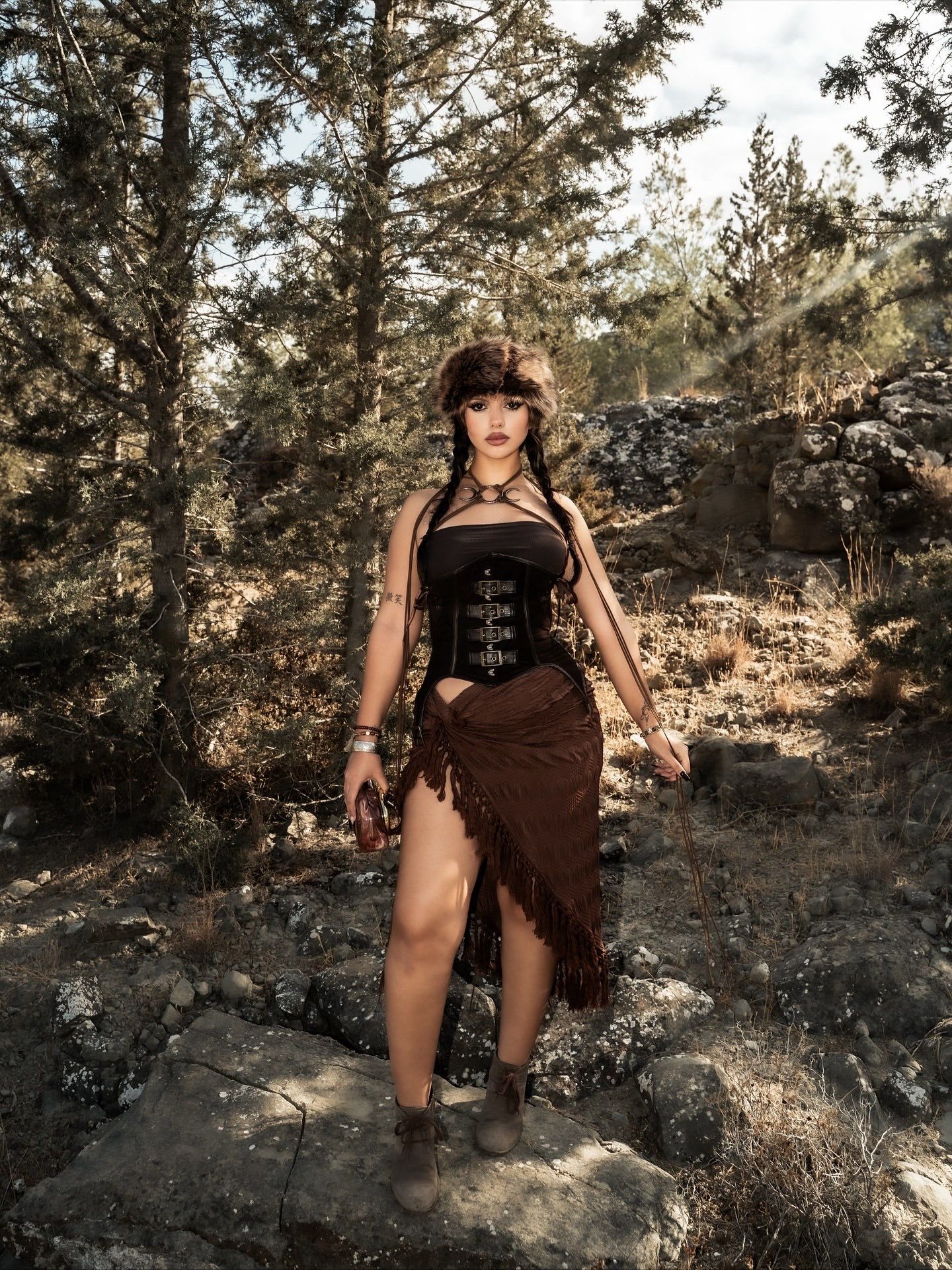 Woman in black corset and brown fringe skirt poses on rocky terrain surrounded by pine trees.